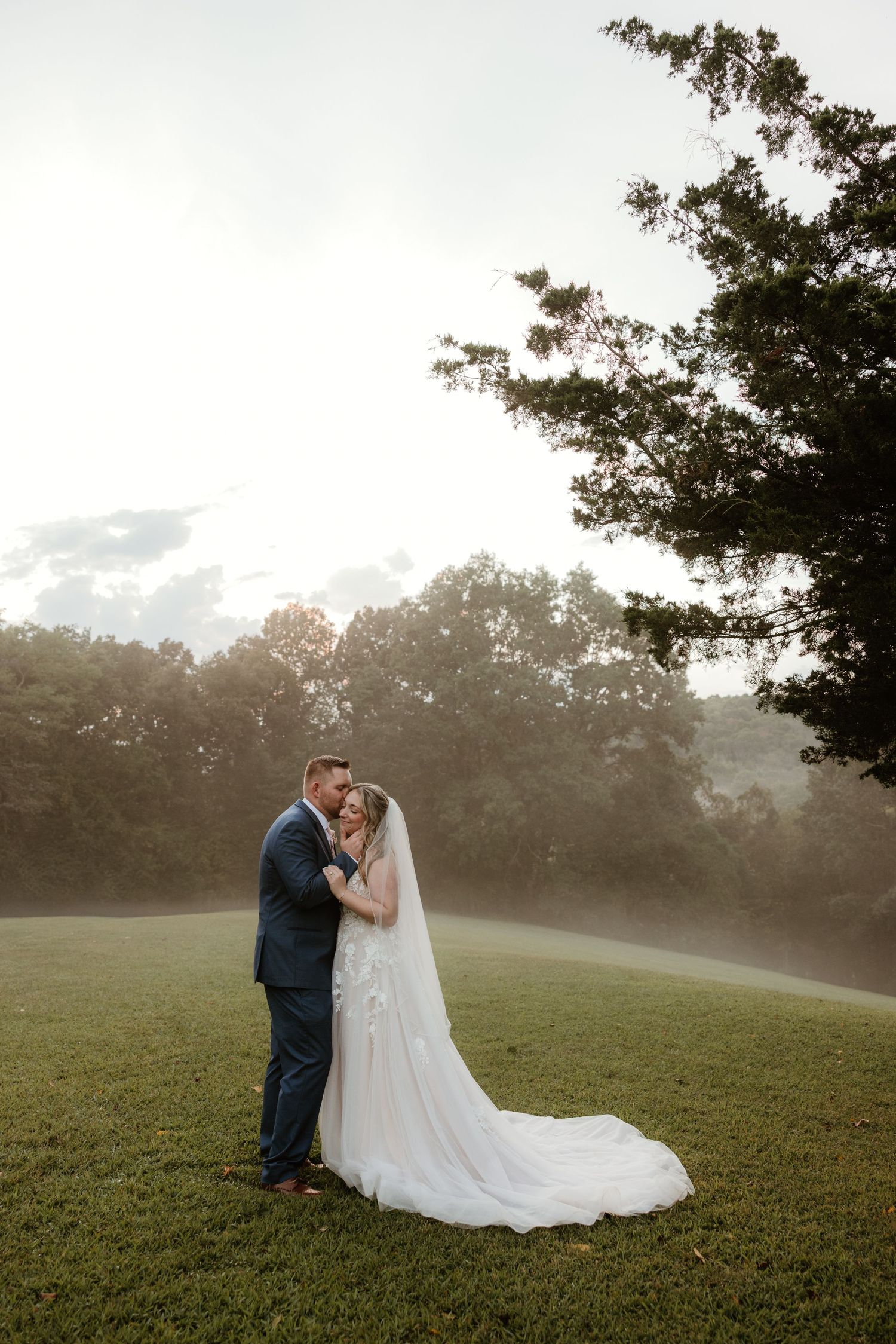 A romantic wedding couple embraces in a misty meadow at sunset with flowing bridal veil and navy suit.