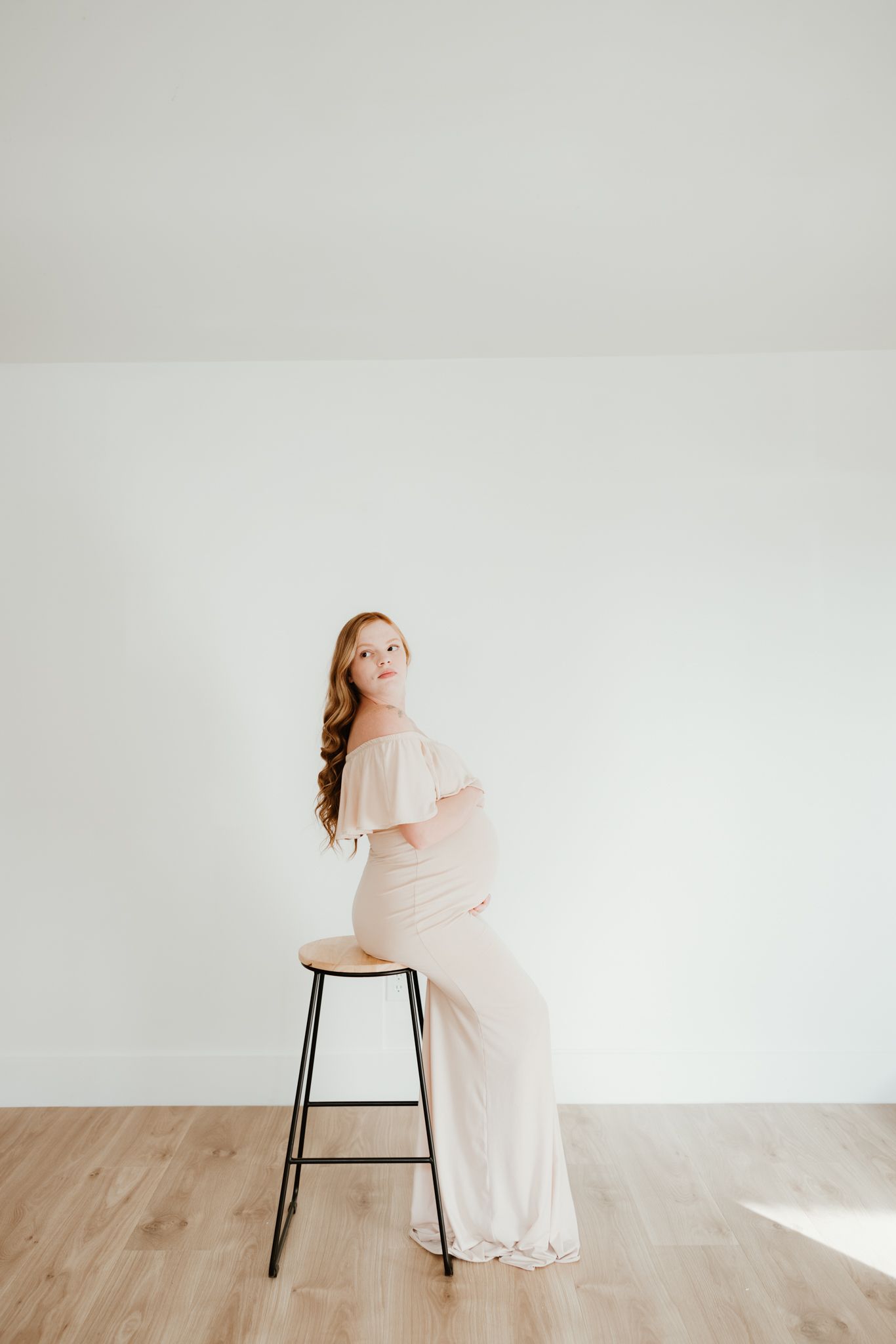 A person in flowing dress poses on stool against white wall in minimalist setting.