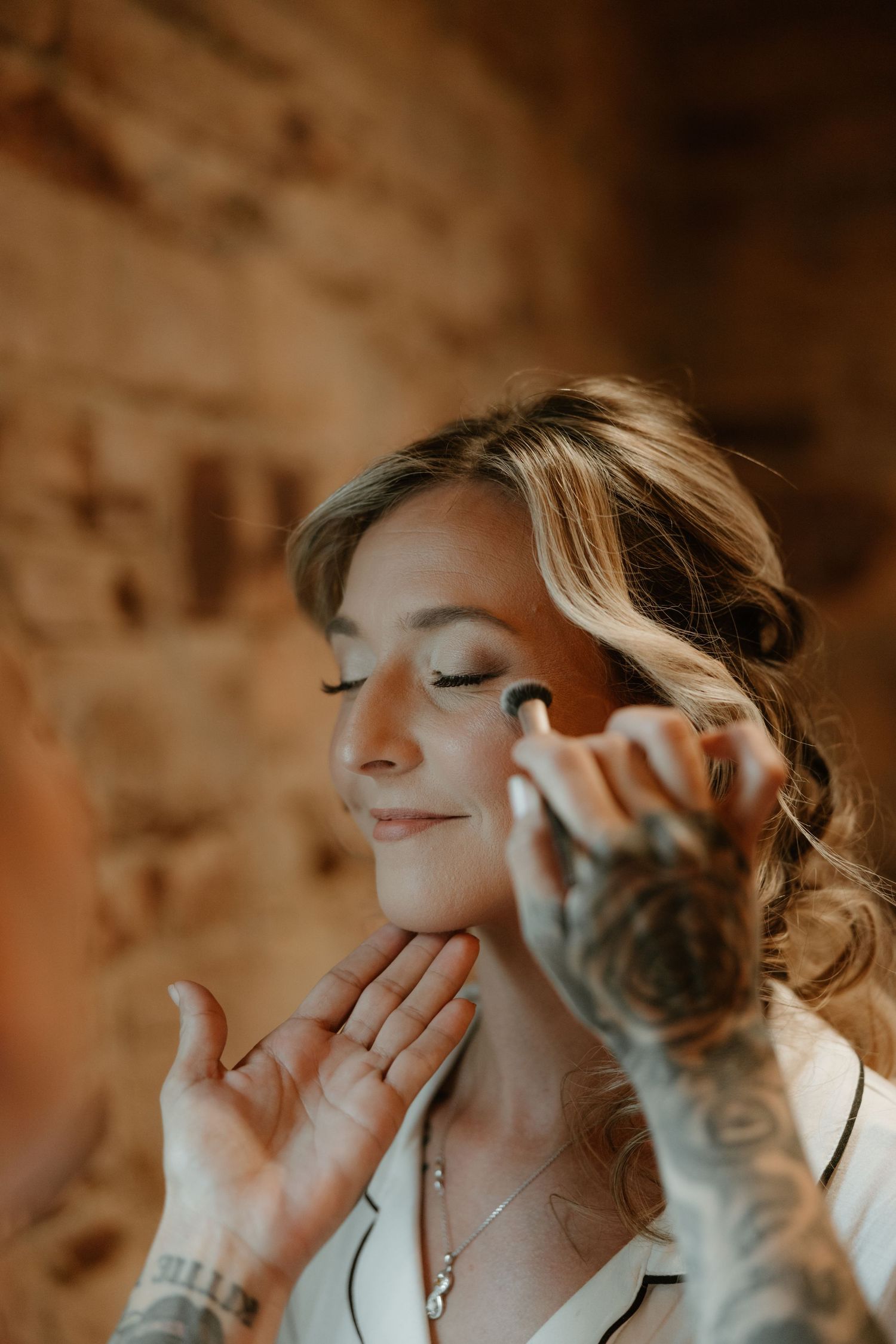 Getting ready photo showing makeup application in warm lighting against stone wall backdrop.