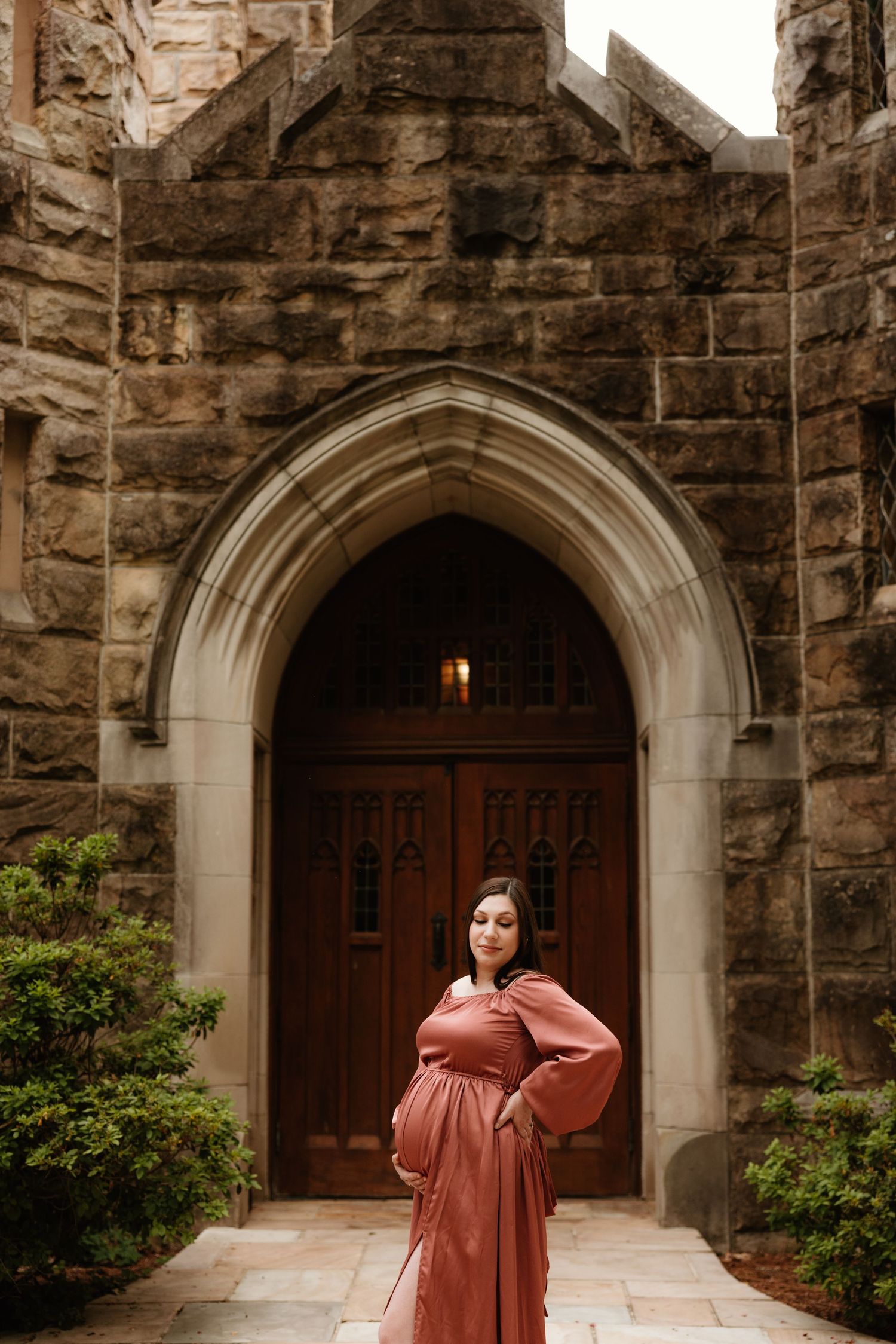 A maternity photo of someone in a rust-colored dress standing in front of a stone church entrance.