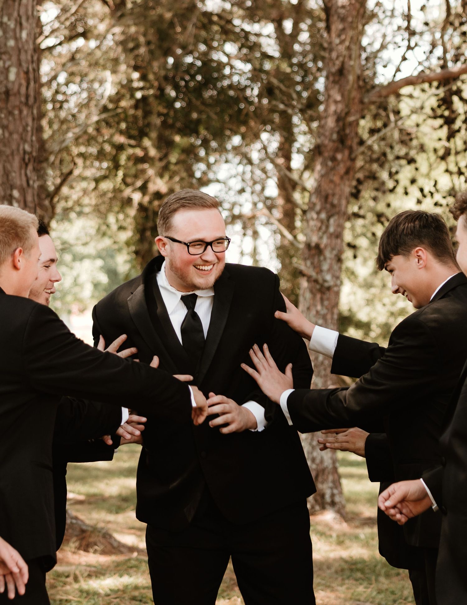 Three groomsmen share a lighthearted moment in black suits among trees during a wedding photoshoot.