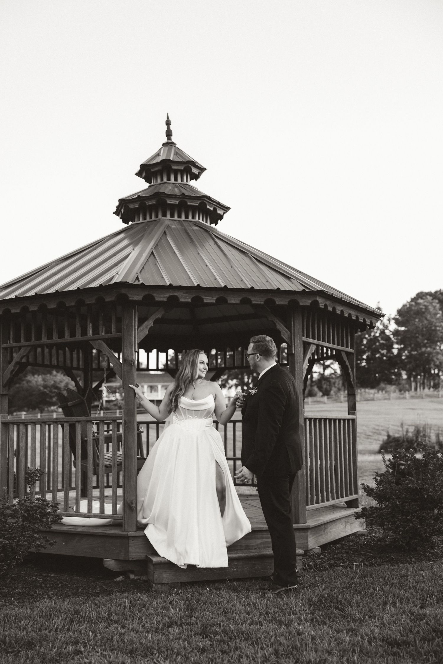 A romantic black and white wedding portrait showing a couple standing in an ornate garden gazebo.