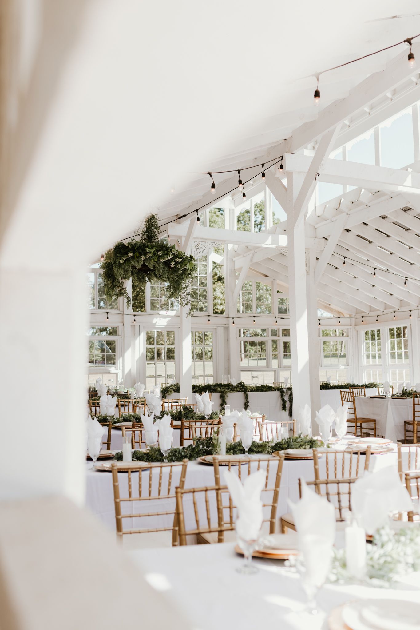 A bright white wedding reception venue with string lights, wooden chairs, and greenery decorations in natural light.