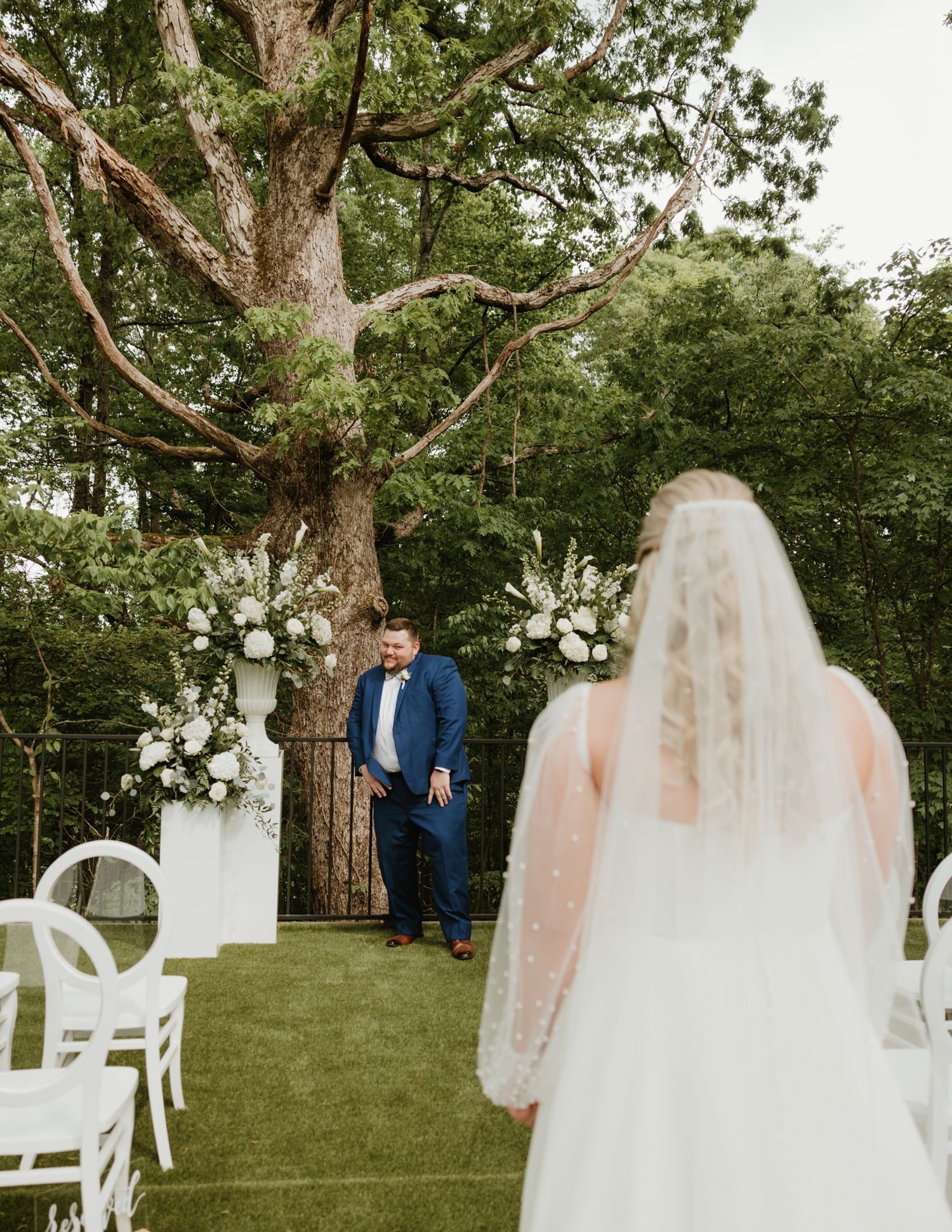 Outdoor wedding ceremony setup with white chairs arranged on a lawn surrounded by mature trees.