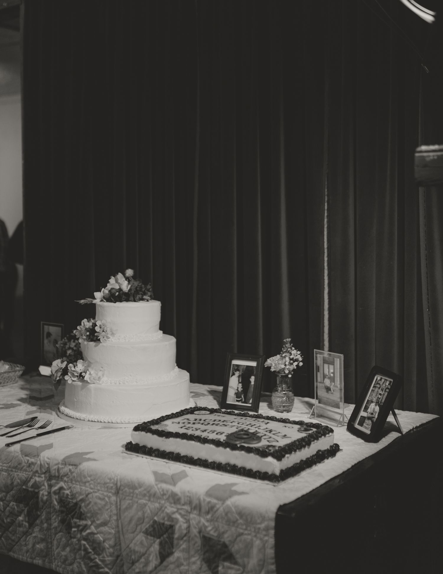 A black and white photo of a wedding cake table with two cakes and framed photographs on display at a reception.