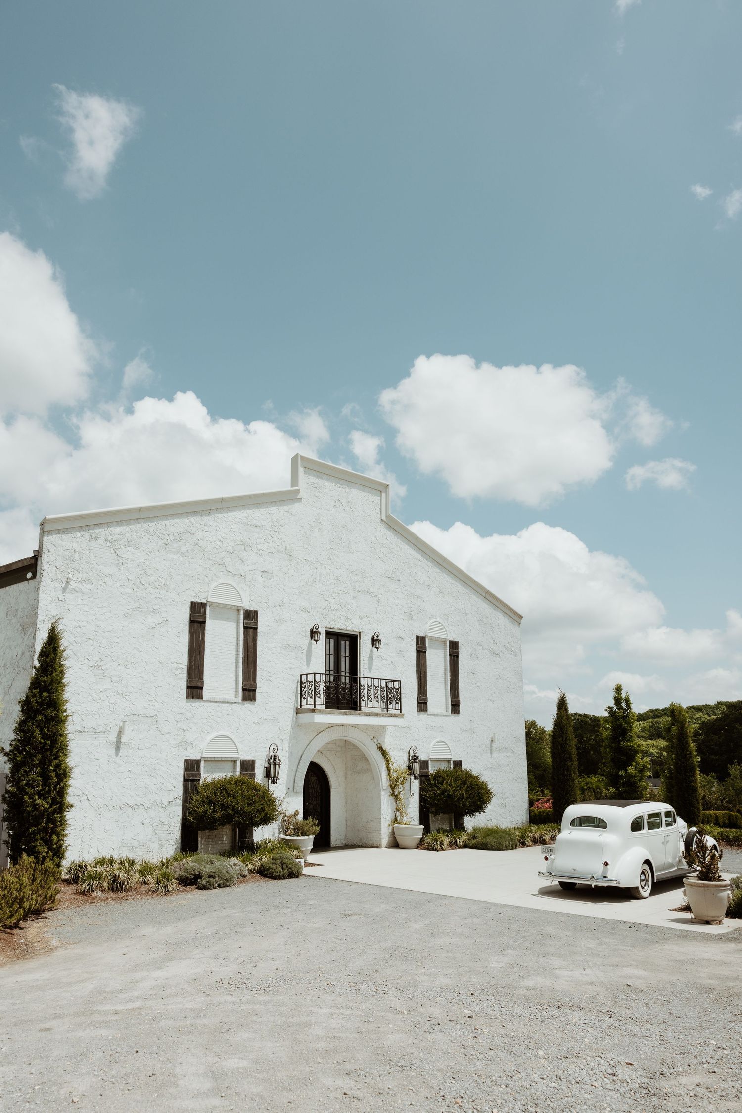 White European-style villa with vintage car parked outside, surrounded by cypress trees under a blue sky.
