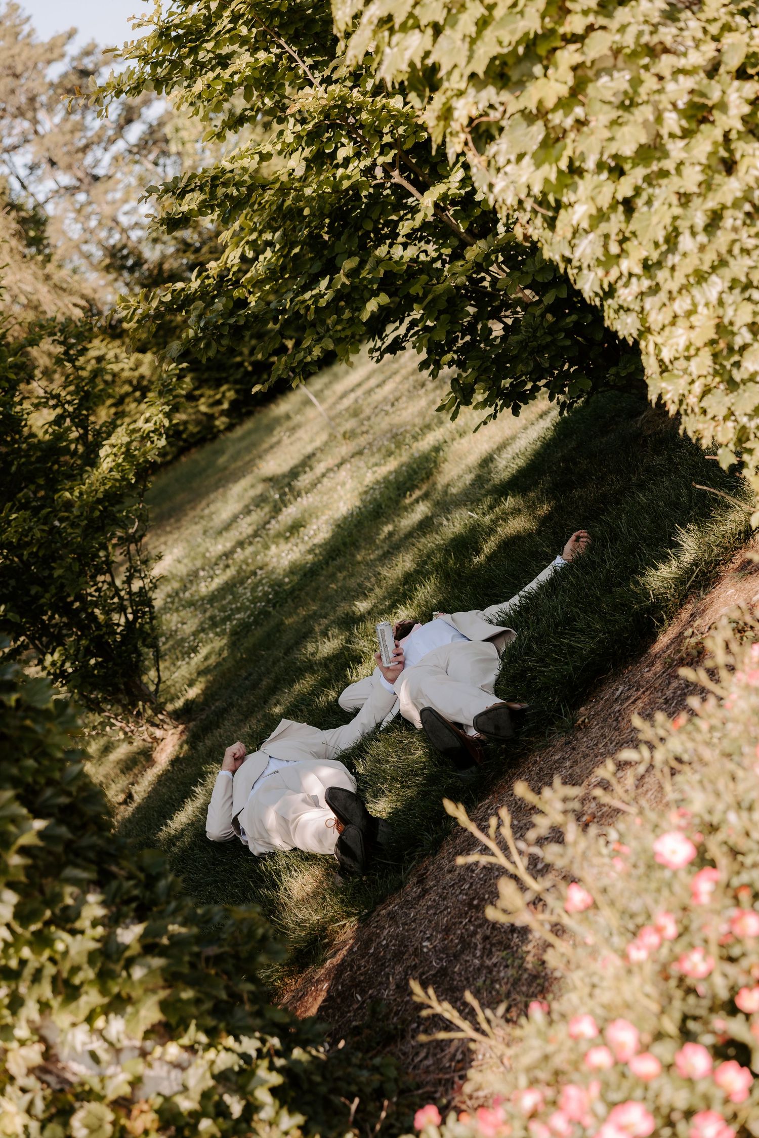 Aerial view of bride and groom lying on lush green grass surrounded by trees and pink flowers.