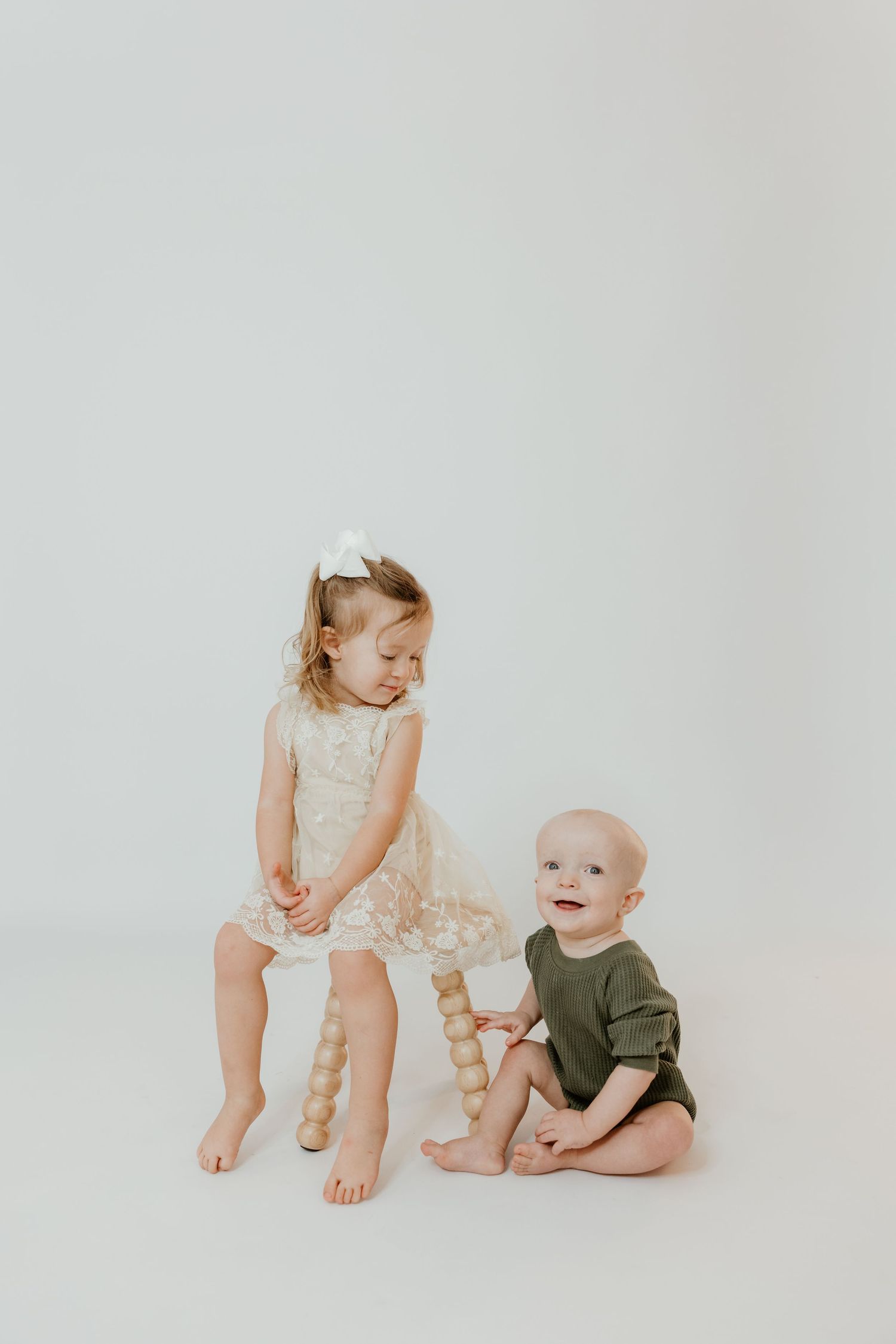 A toddler girl in a lace dress sits on a wooden stool beside a smiling baby boy in a green romper on a white background.