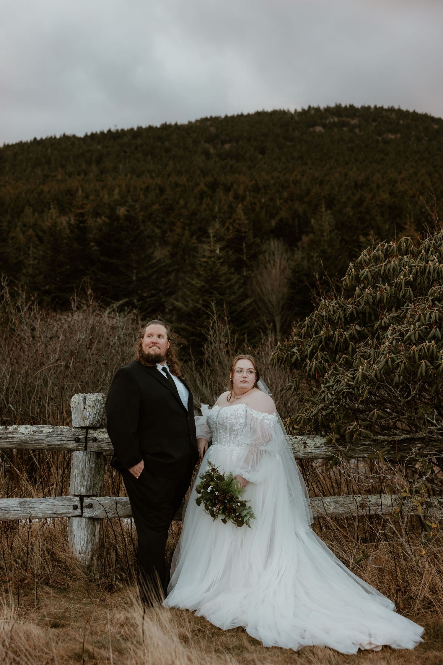 Wedding couple poses by a rustic fence with mountain backdrop, bride in white tulle gown holding greenery bouquet.