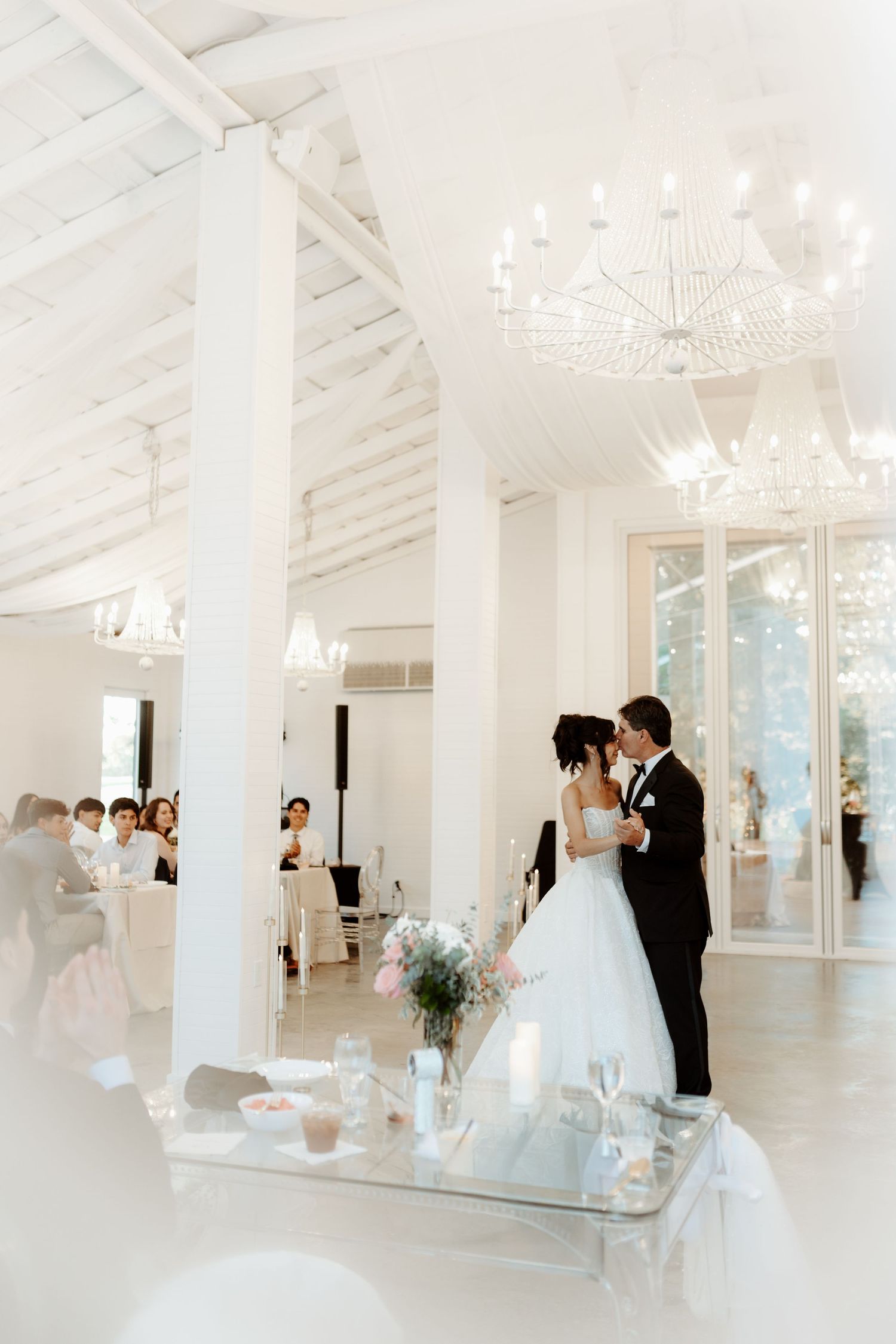 Bride and groom share first dance in elegant white ballroom with crystal chandeliers and floral centerpieces at wedding reception.