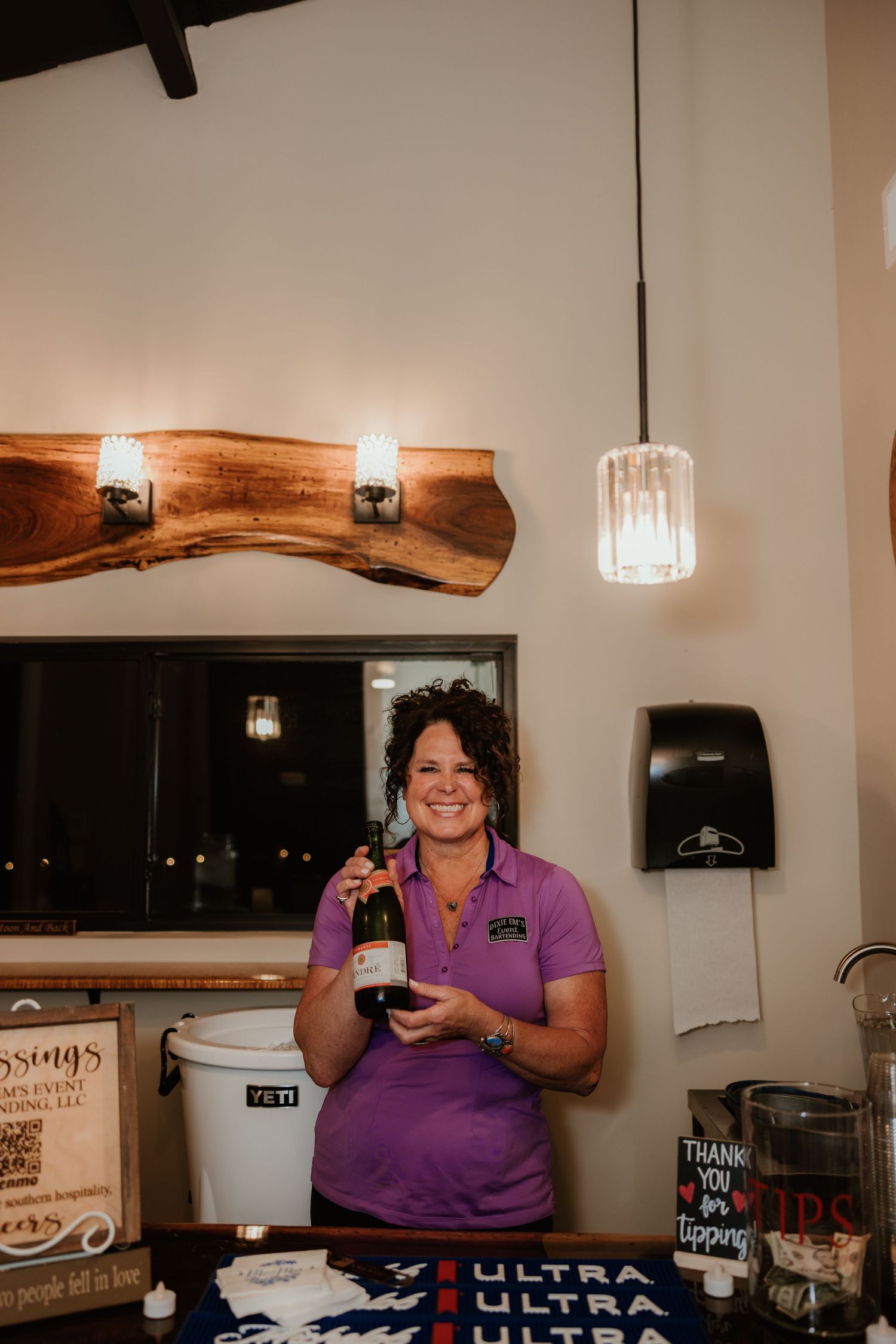 A bartender in purple shirt stands behind a counter with drink supplies and illuminated by warm rustic wood lighting fixtures.