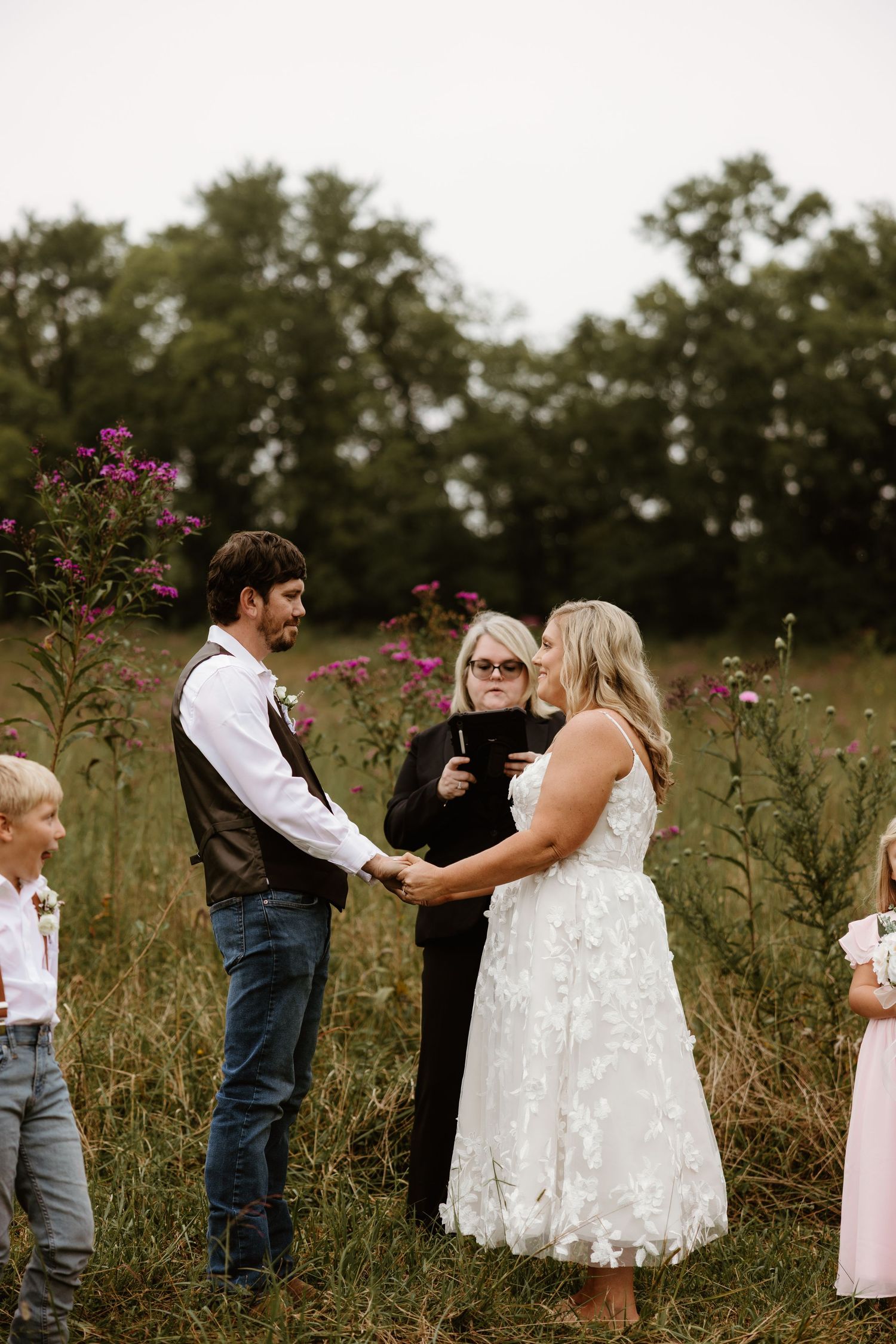 A wedding ceremony takes place in a rustic field with wildflowers and tall grass on an overcast day.