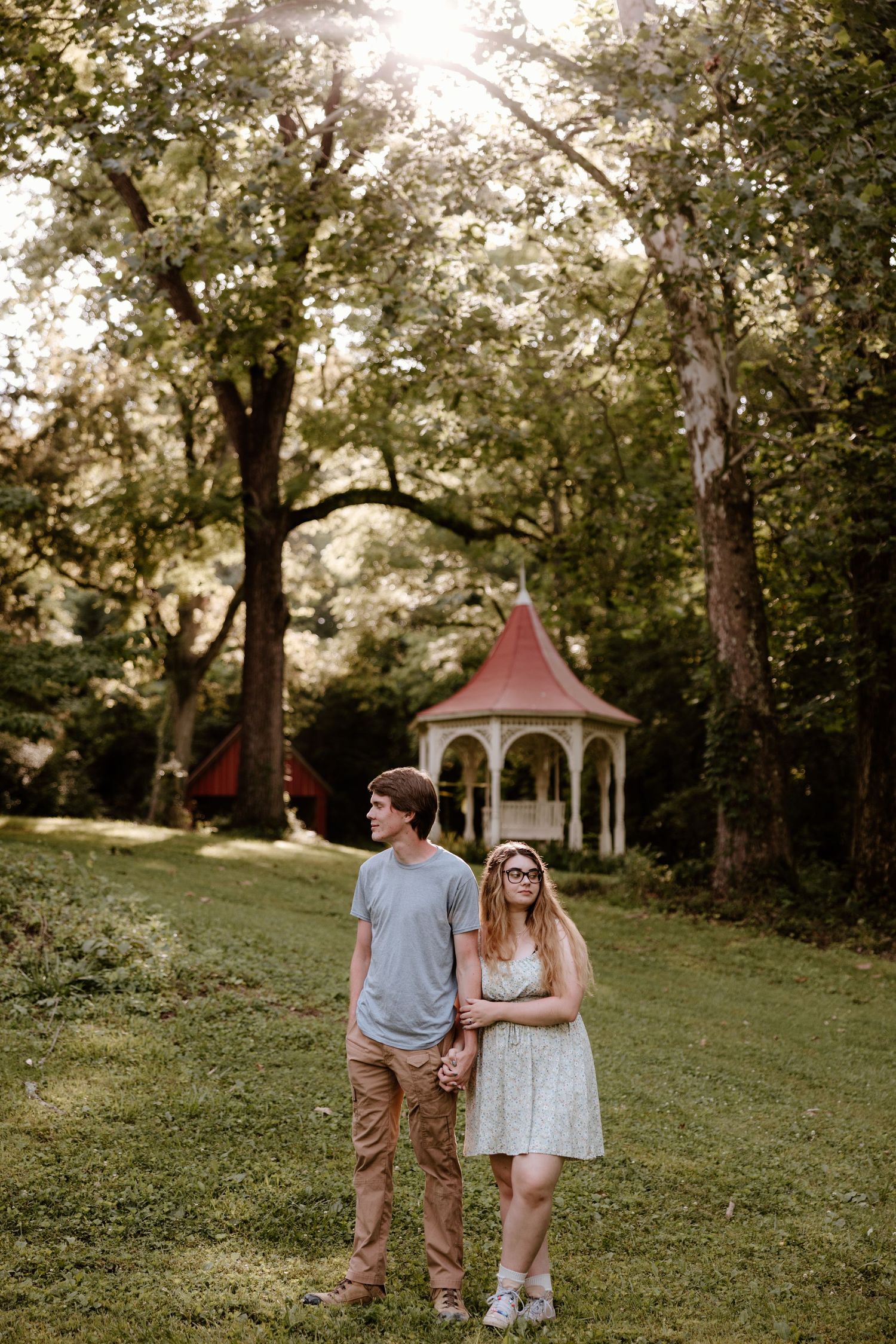 A couple stands in a garden with a white gazebo and trees in the background during golden hour.