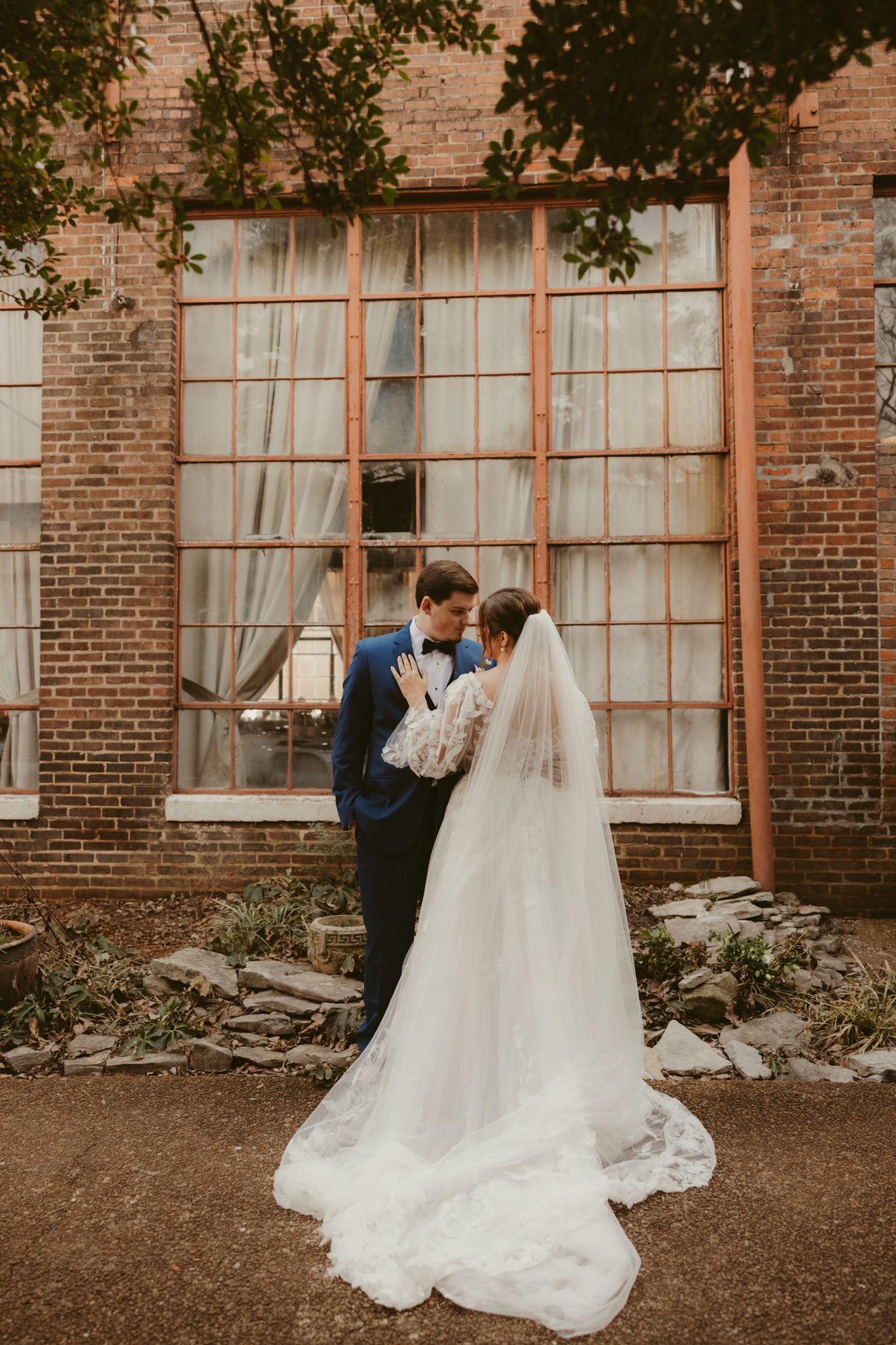 A romantic wedding portrait against a rustic brick wall with large industrial windows.