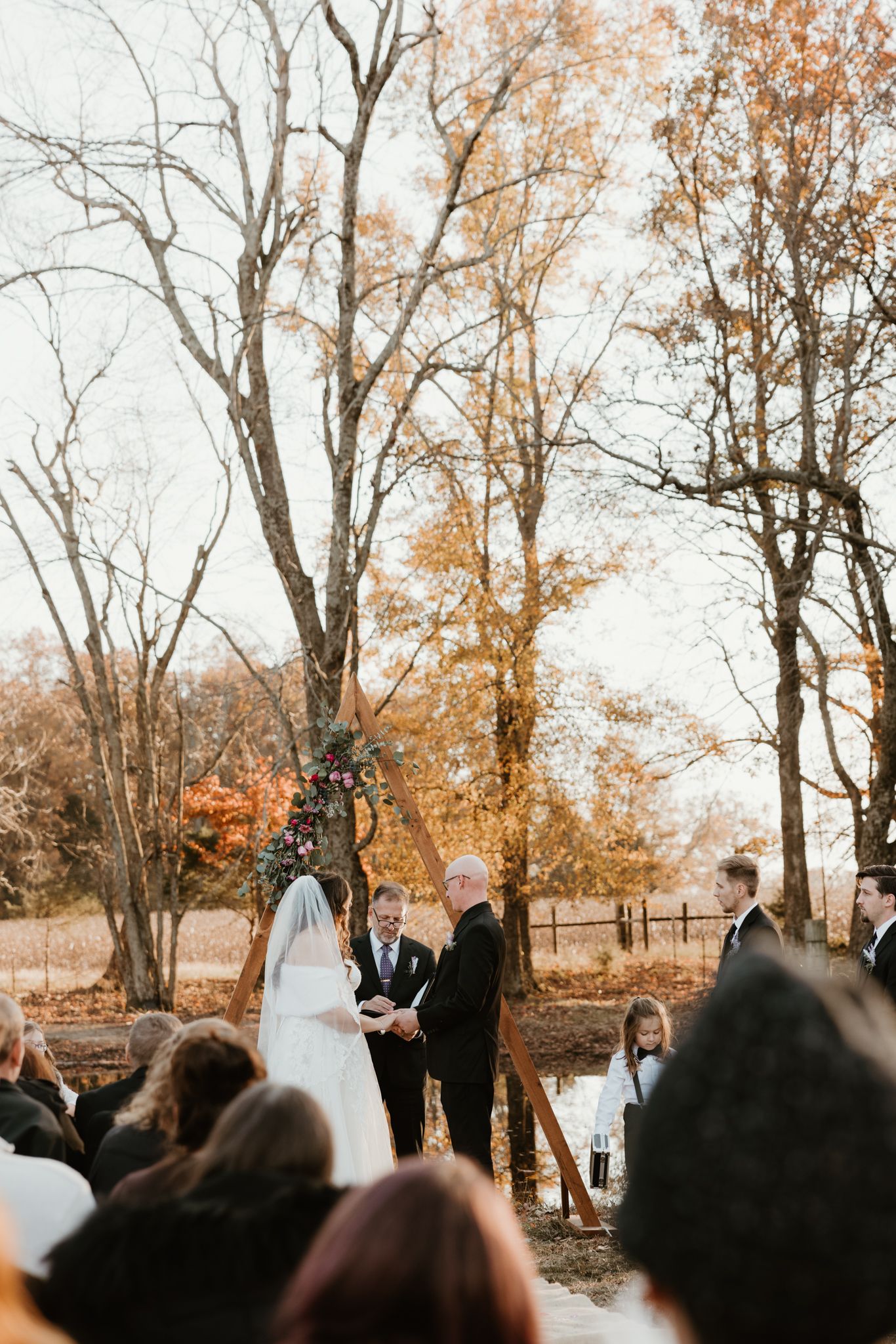An outdoor autumn wedding ceremony surrounded by golden trees and gathered guests.