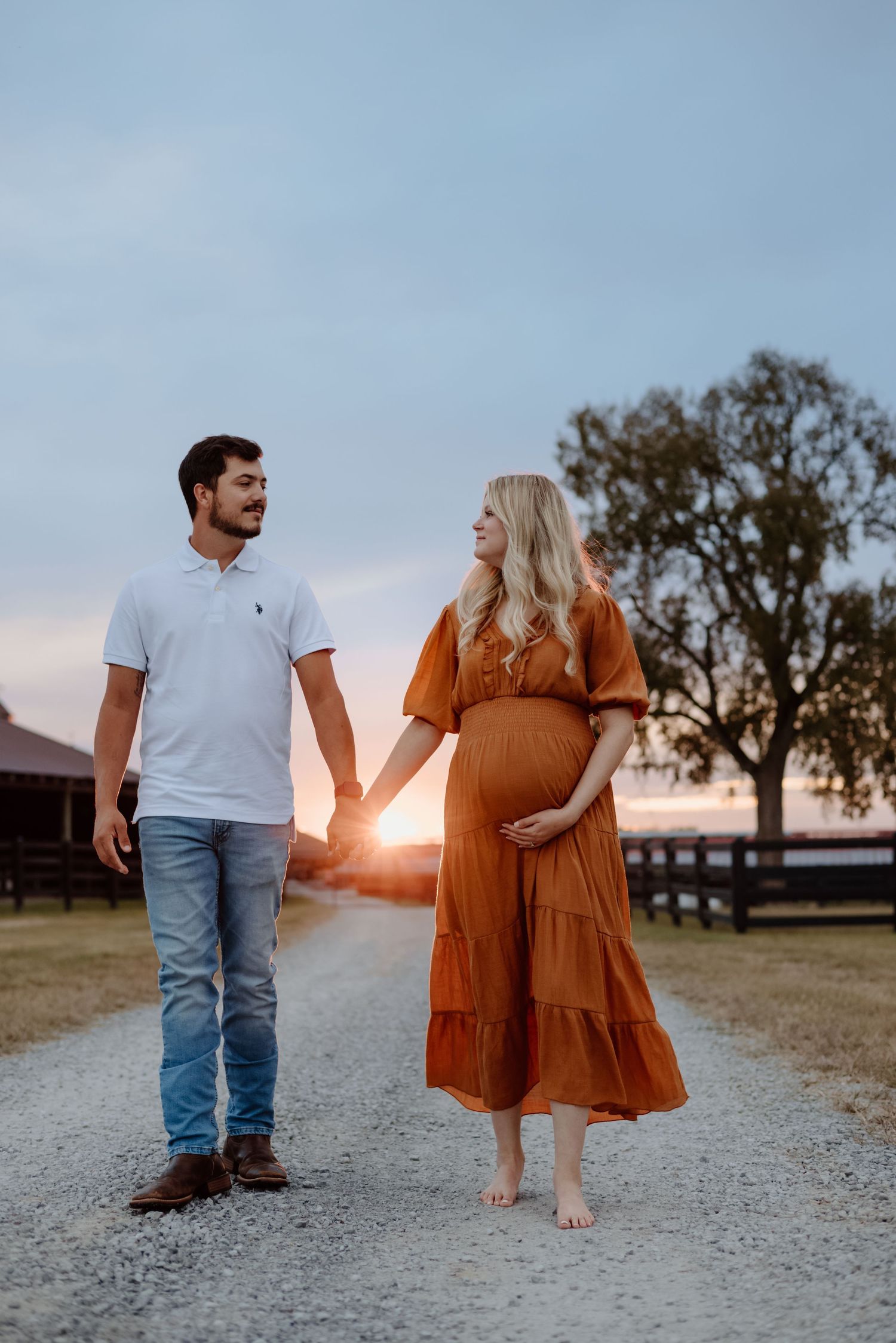 A romantic couple stands together at sunset on a gravel path, with one wearing a rust-colored dress and the other in jeans.