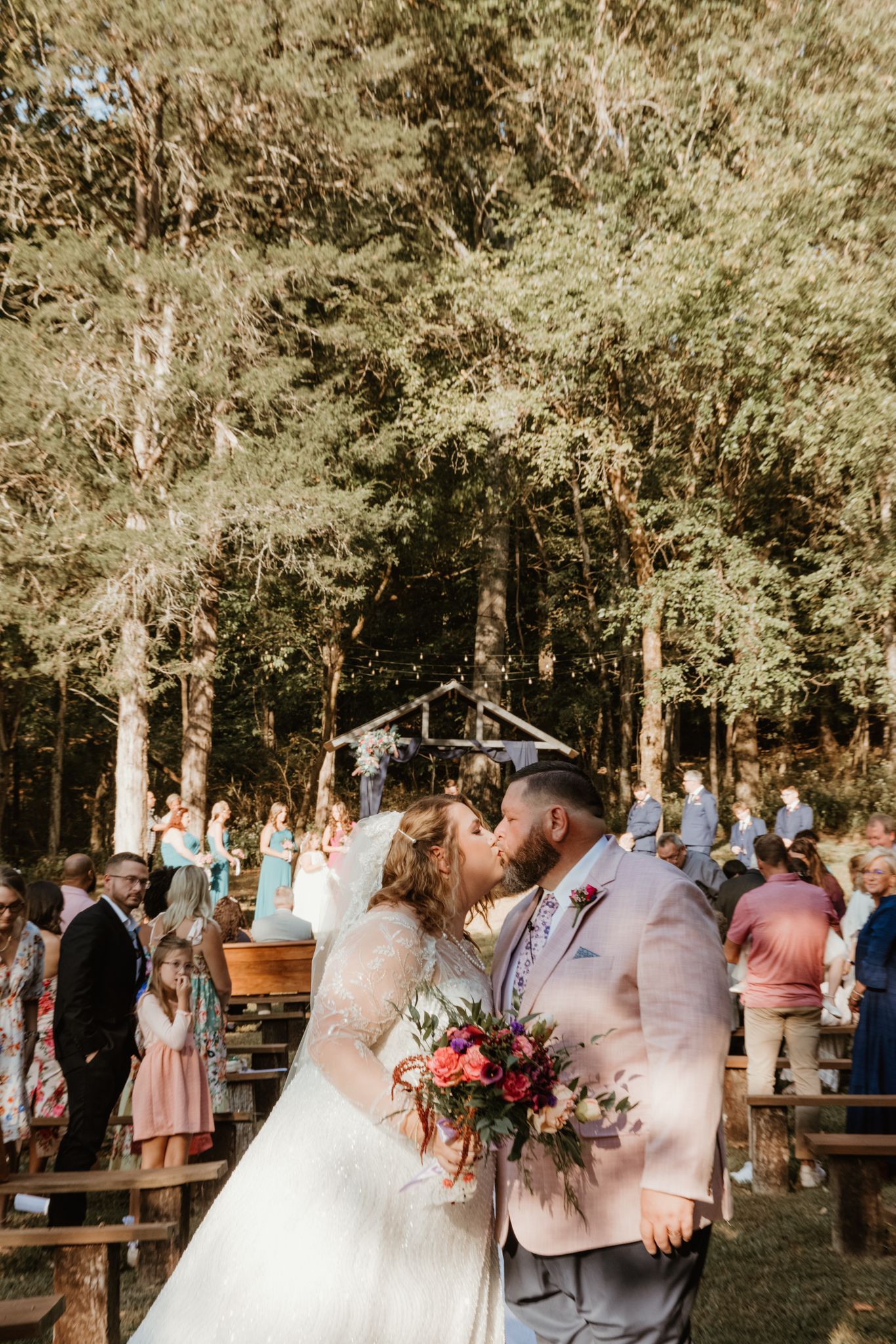 A romantic outdoor wedding ceremony in a wooded area with wooden benches and guests gathered under trees.