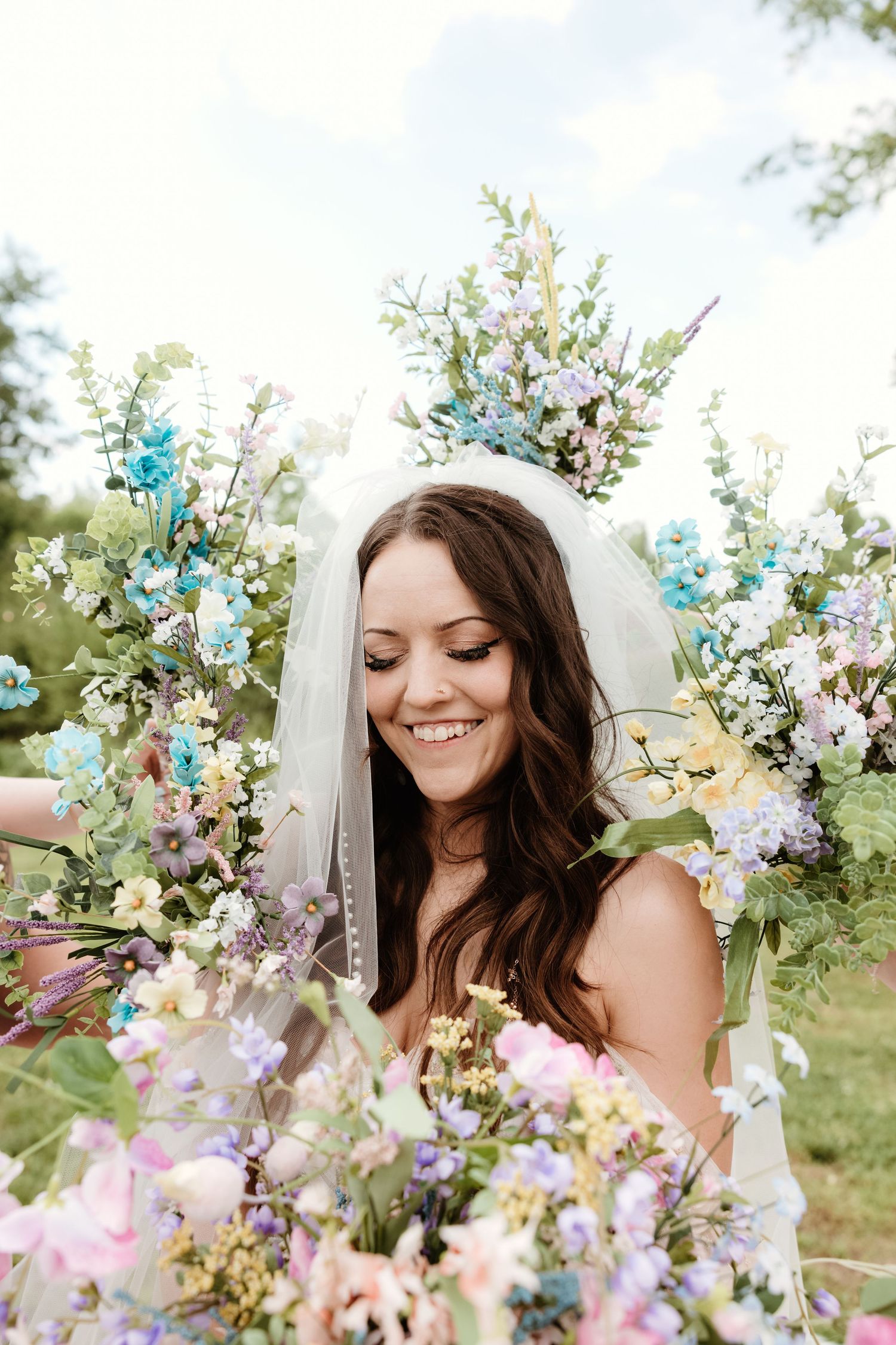 A bride adorned in white glows with joy amidst a garden filled with pastel blue and purple florals and delicate foliage.