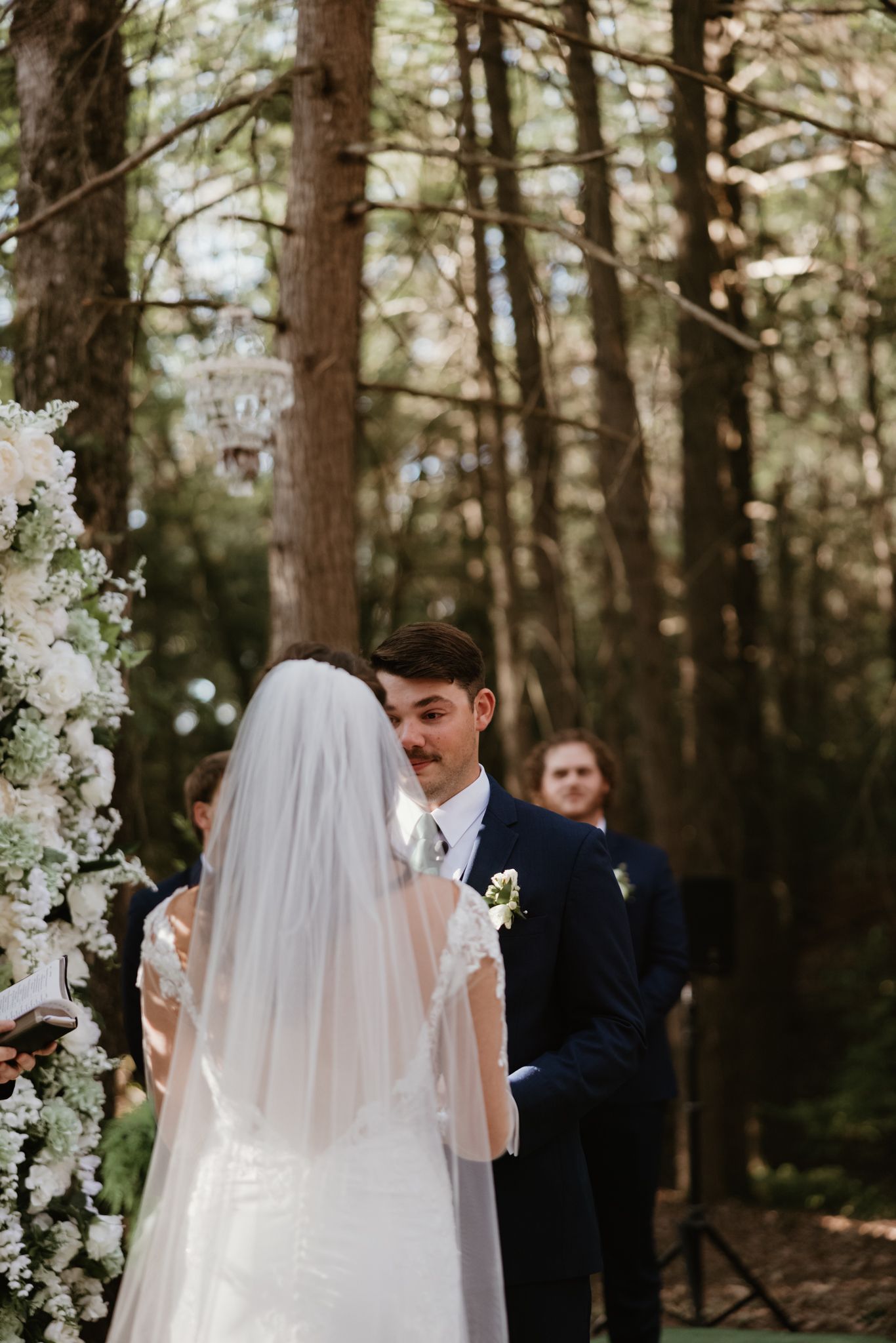 Wedding ceremony in a wooded setting with white floral arch decorations.