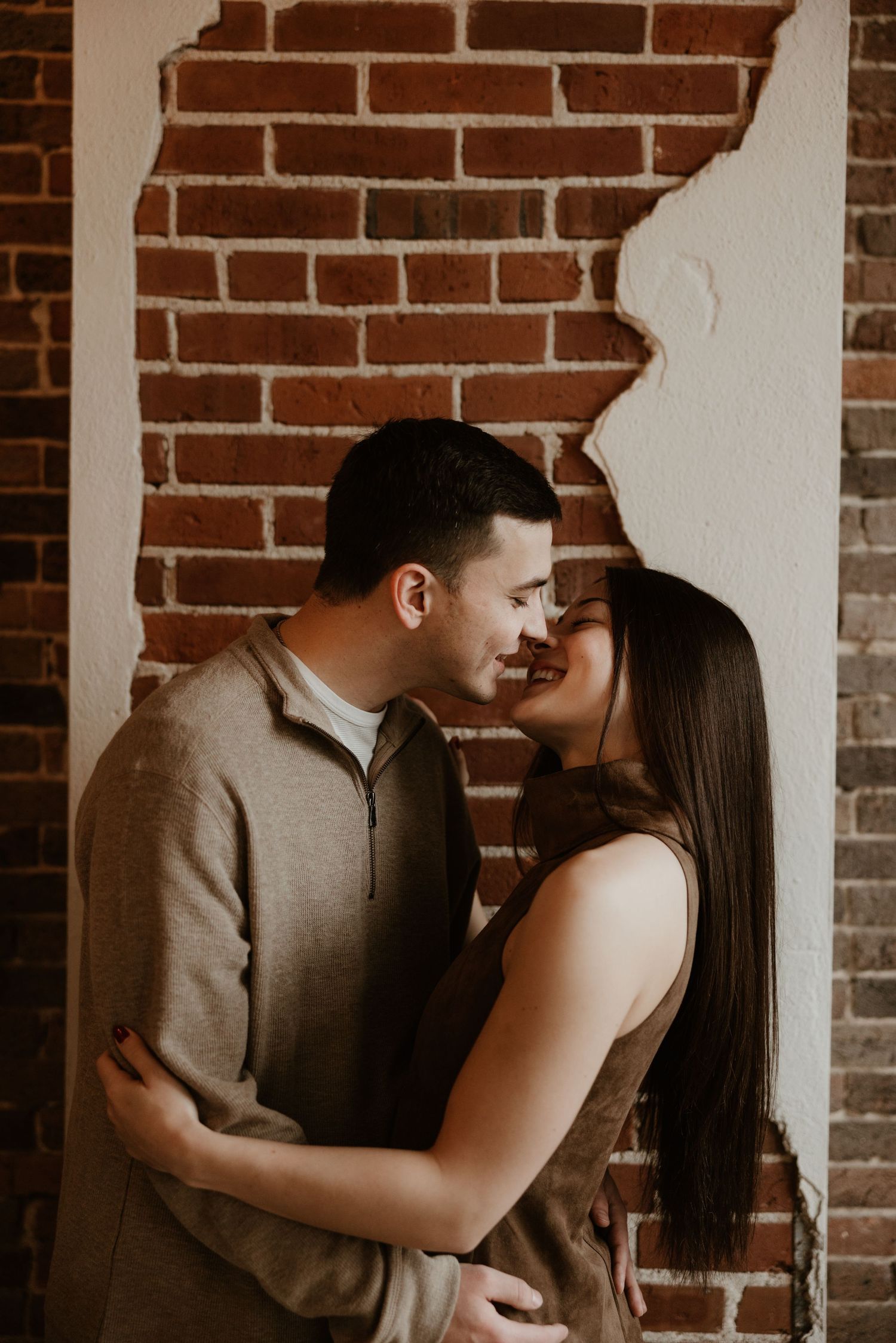 Couple sharing a romantic moment, nearly kissing, embracing against a rustic brick wall in warm, intimate lighting.