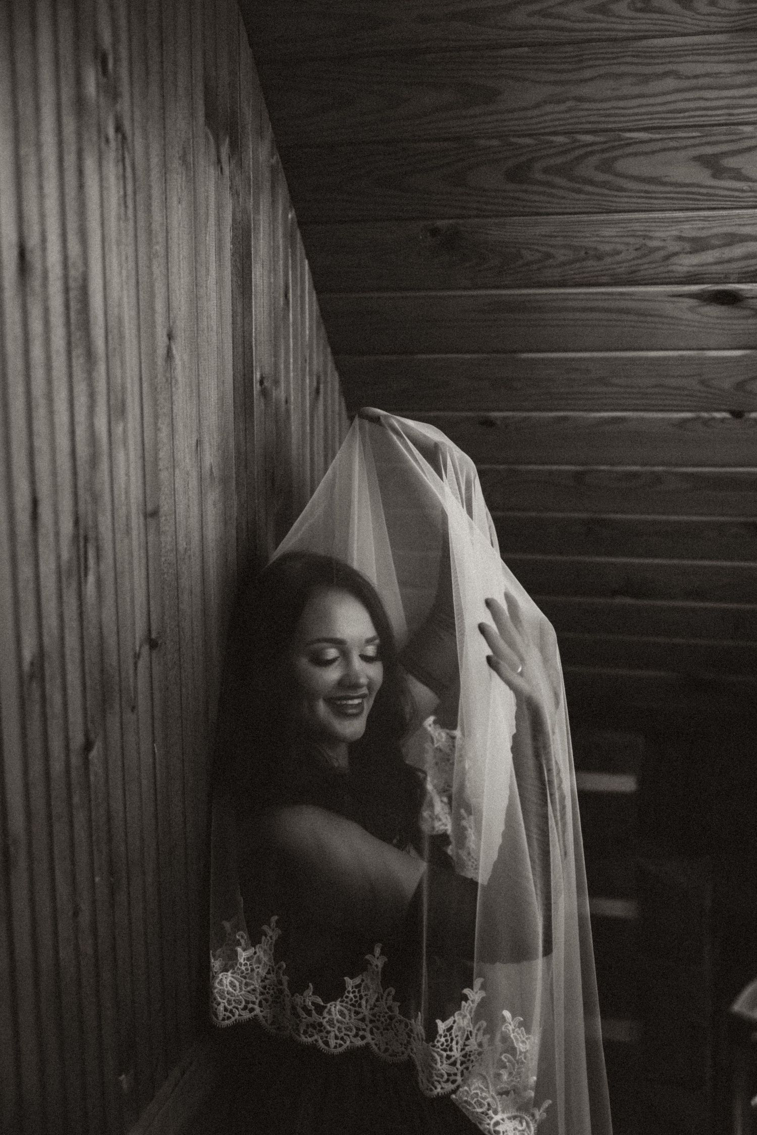 Black and white photo of a smiling bride holding her lace veil over her face in a rustic wooden interior.