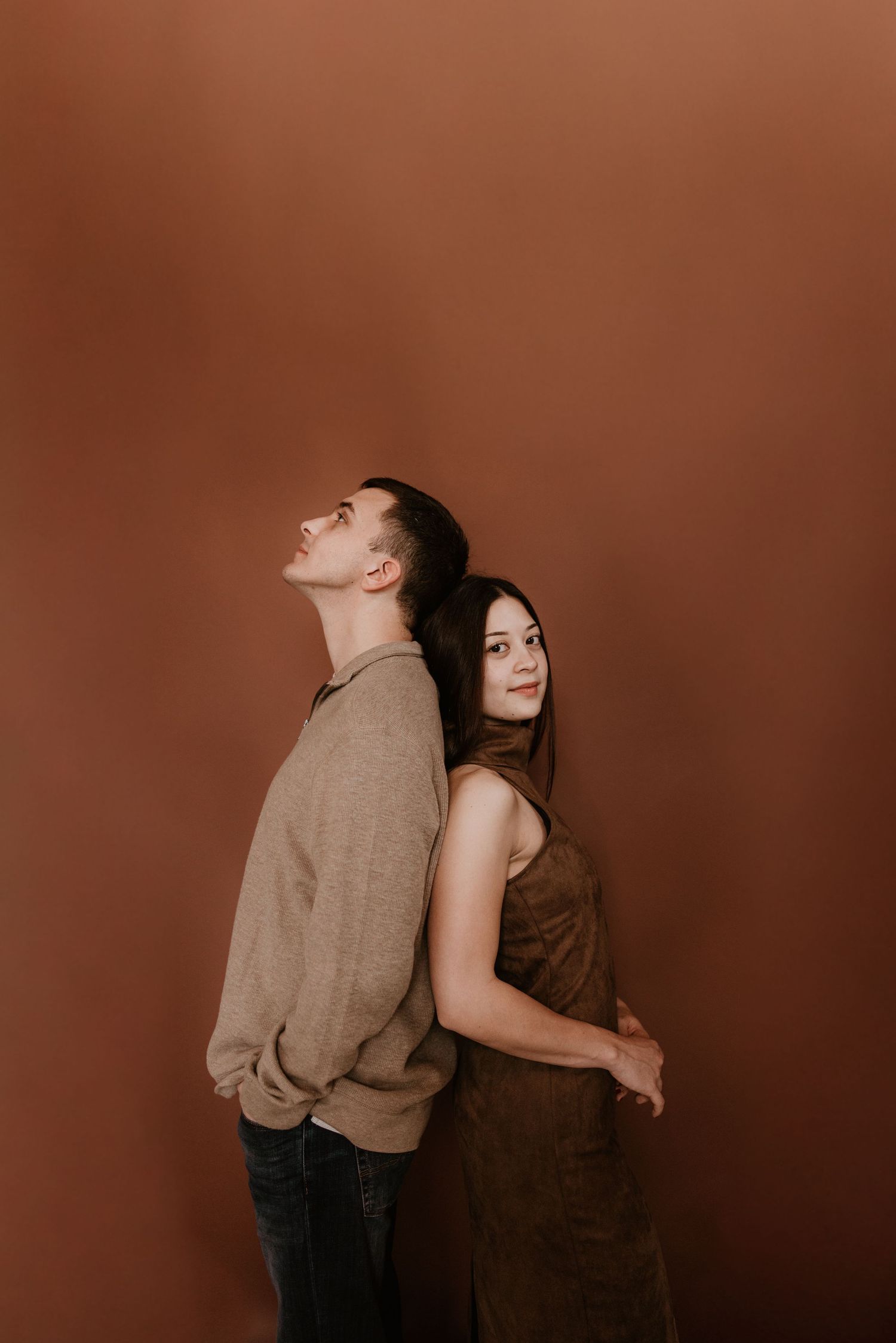 A man and woman stand back-to-back against a brown wall, wearing earth-toned outfits, as she smiles at the camera.