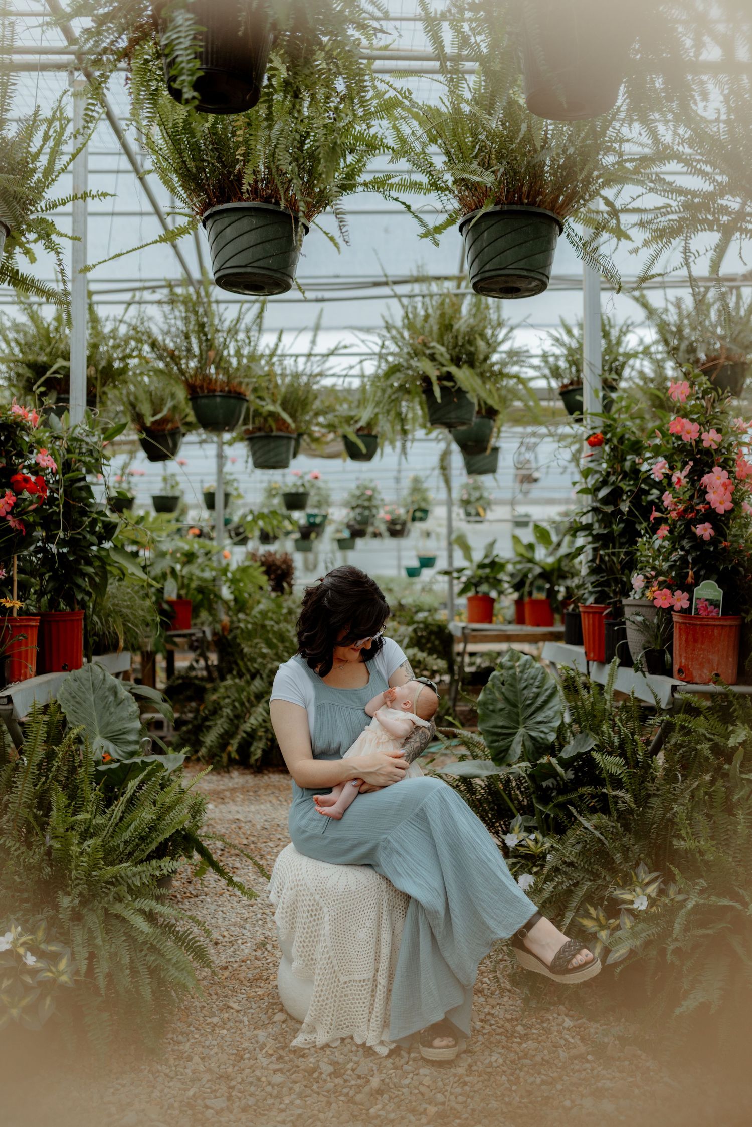 A woman in a blue dress holds a baby while seated in a lush greenhouse surrounded by hanging ferns and potted plants.