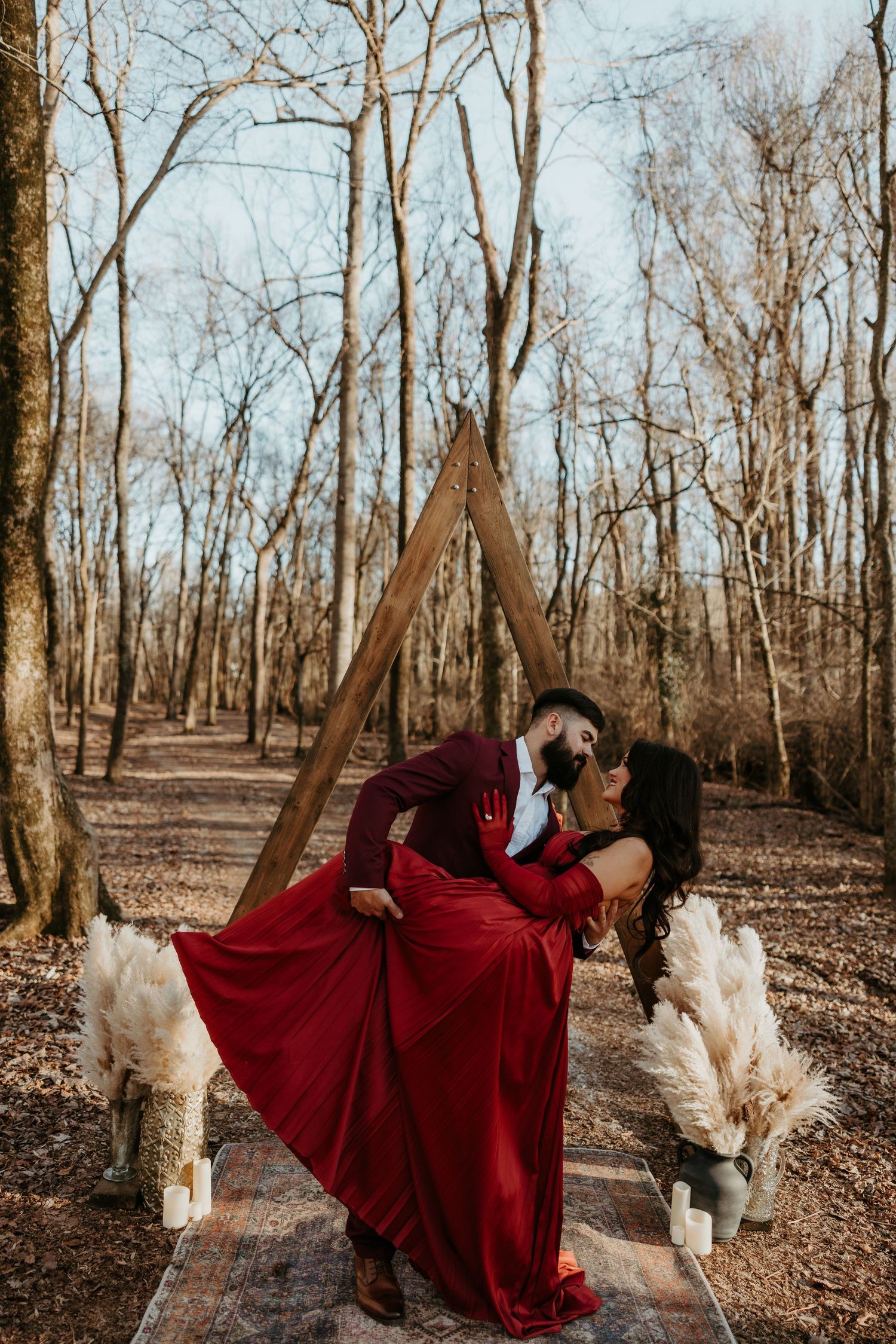 Couple in matching red outfits sharing a romantic kiss under a triangular wooden arch in a forest setting.