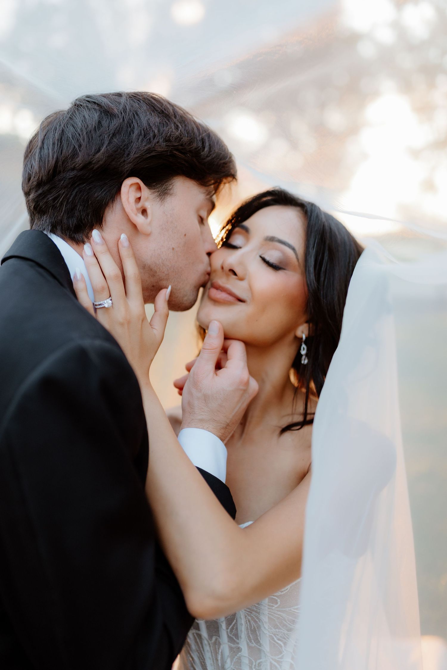 Groom in black tuxedo tenderly kisses bride in white feathered dress under veil during romantic wedding portrait.