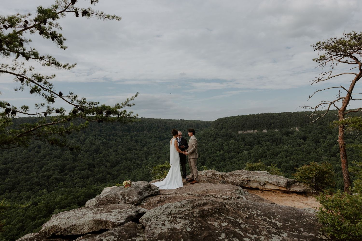 Couple exchanges vows on a rocky mountain overlook surrounded by lush green forest during an outdoor wedding ceremony.