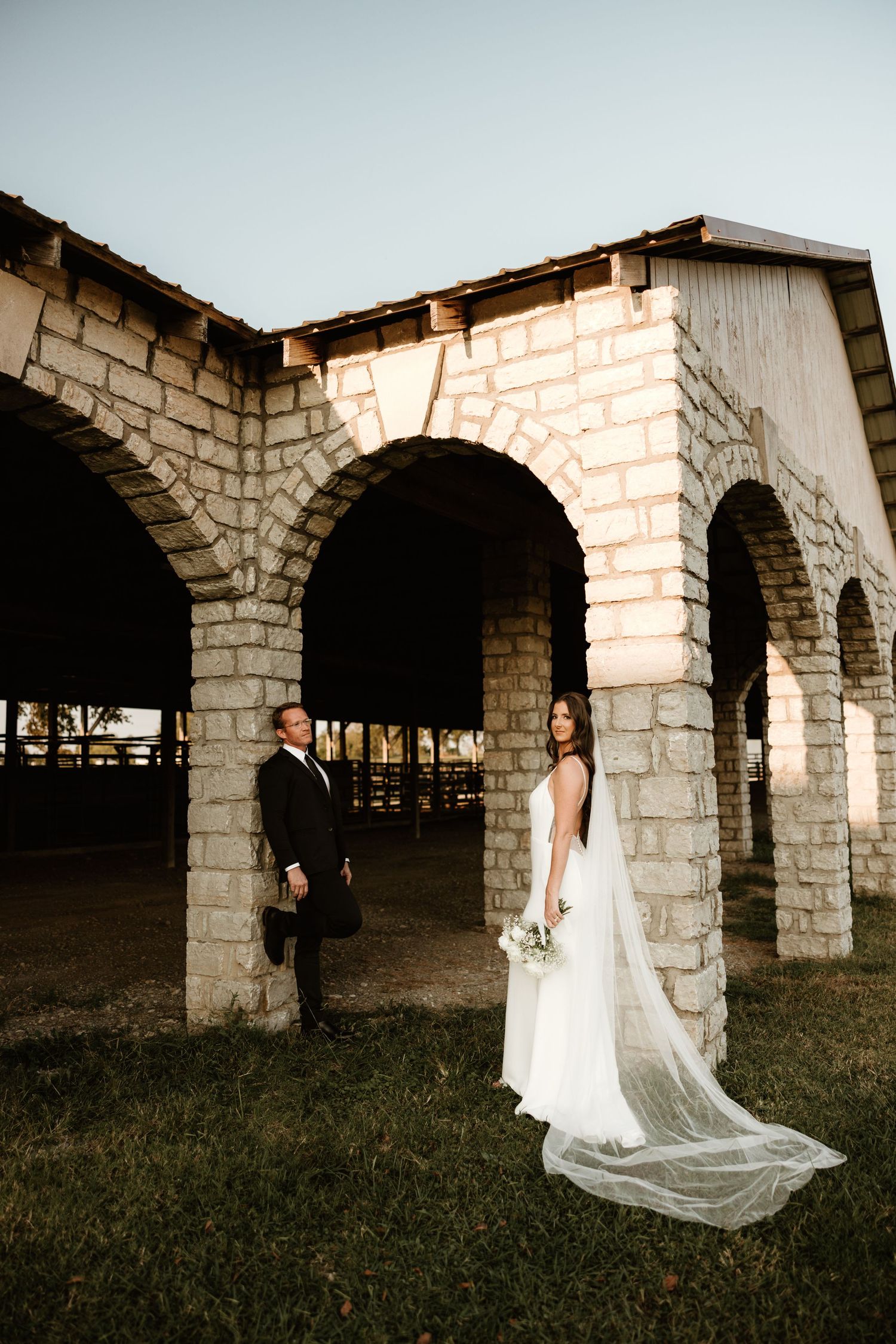A rustic stone archway provides a romantic backdrop for a flowing white wedding dress against the natural setting at sunset.