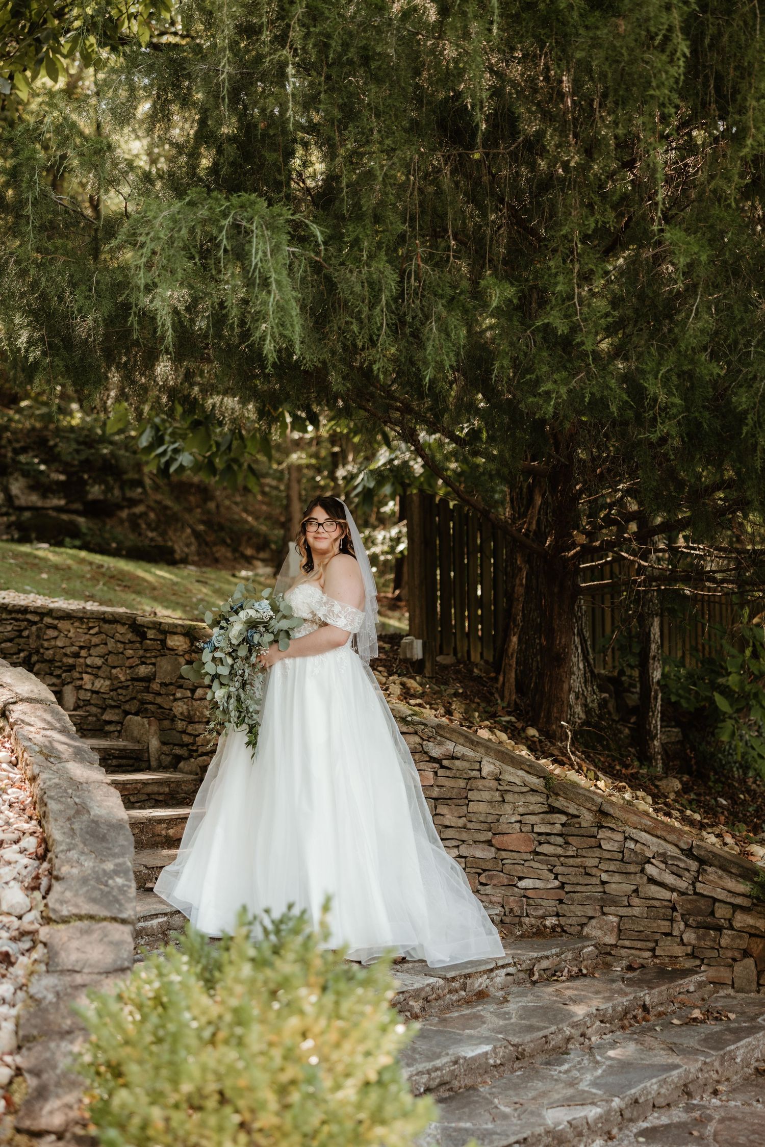 A bride in a flowing white ballgown poses on stone steps surrounded by lush greenery at an outdoor wedding venue.
