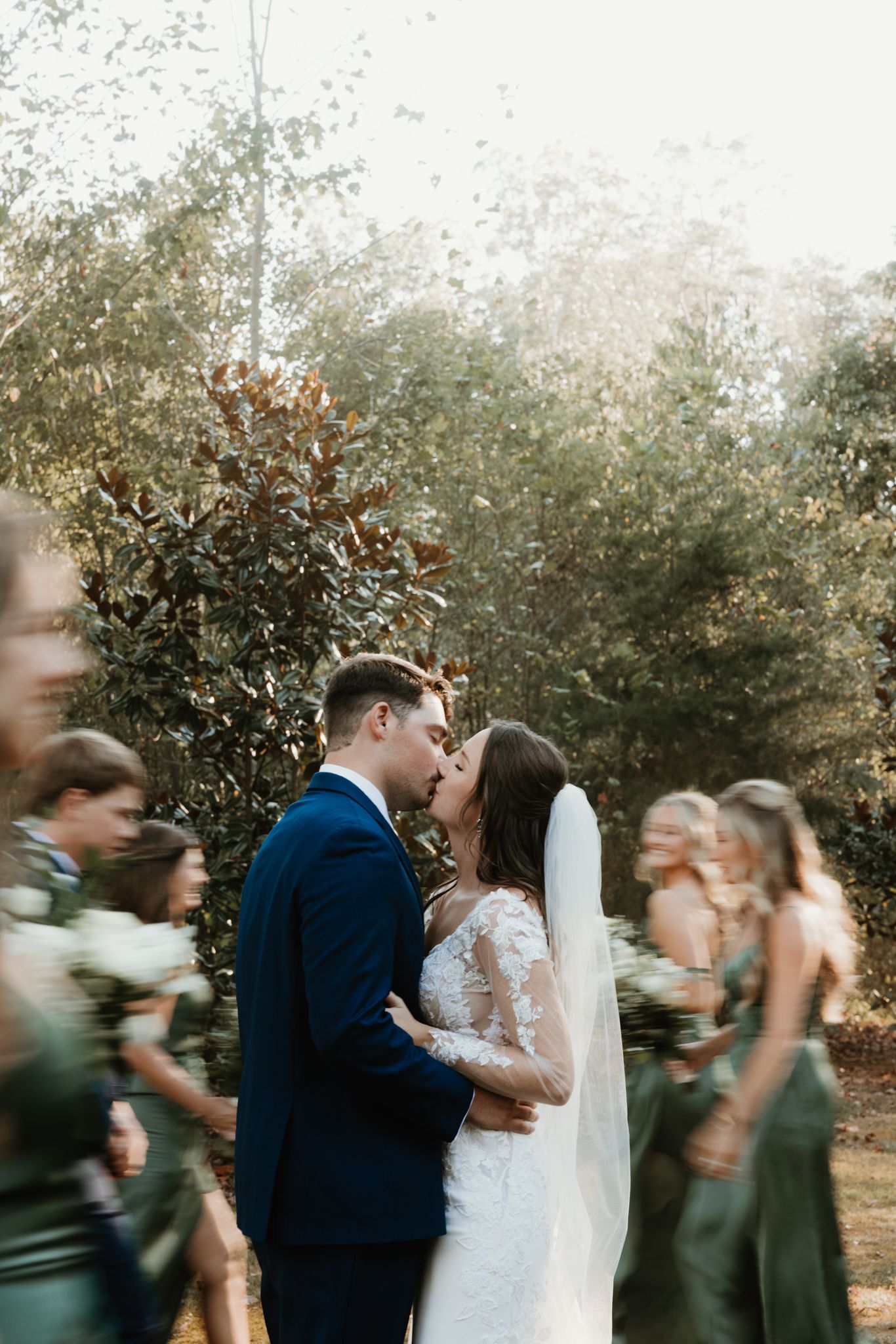 A romantic wedding moment captured outdoors with guests in dark green dresses and natural greenery in the background.