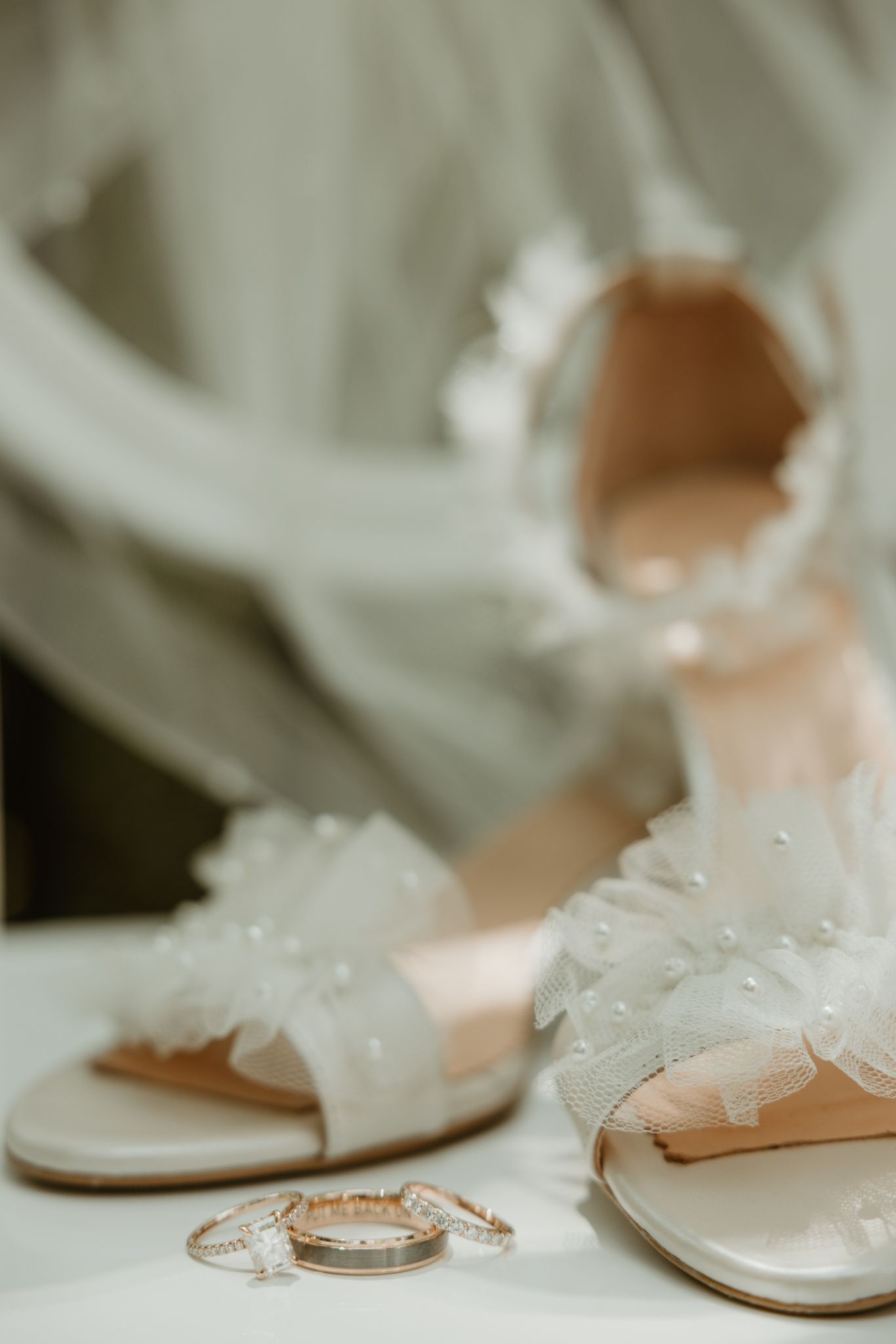 Elegant white bridal shoes with delicate pearl details next to wedding rings on a light surface.