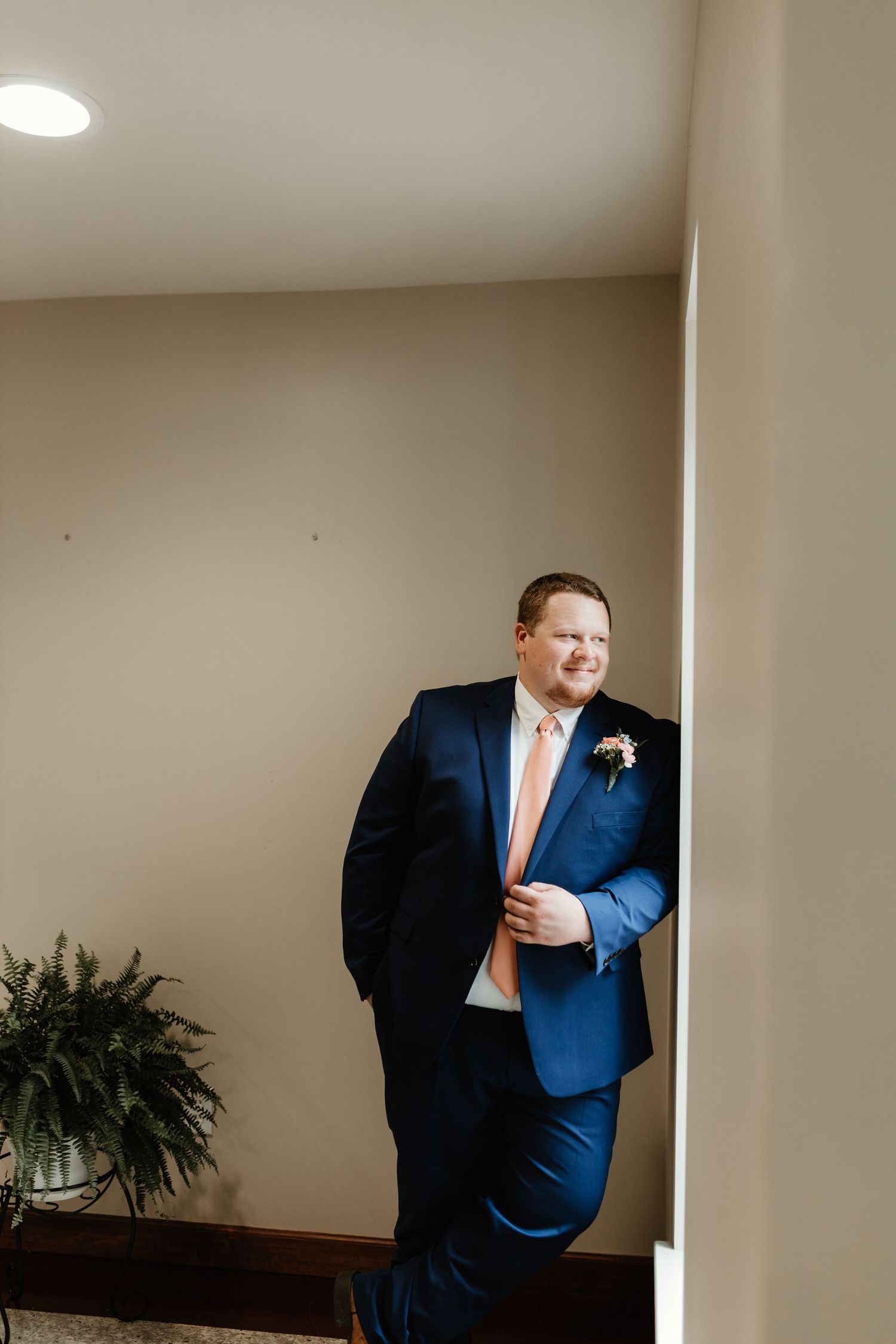 A groom in a navy blue suit and peach tie leans casually against a wall near a decorative fern at an indoor venue.