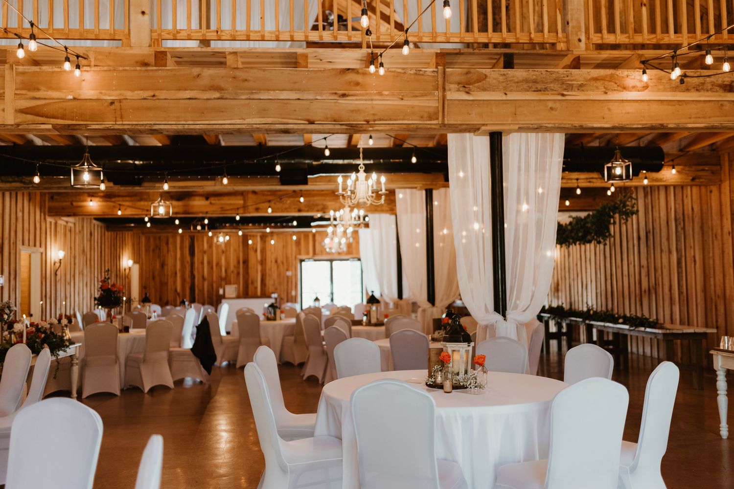 Elegant rustic wedding reception venue with white tablecloths, wooden walls, and hanging string lights creating warm ambiance.