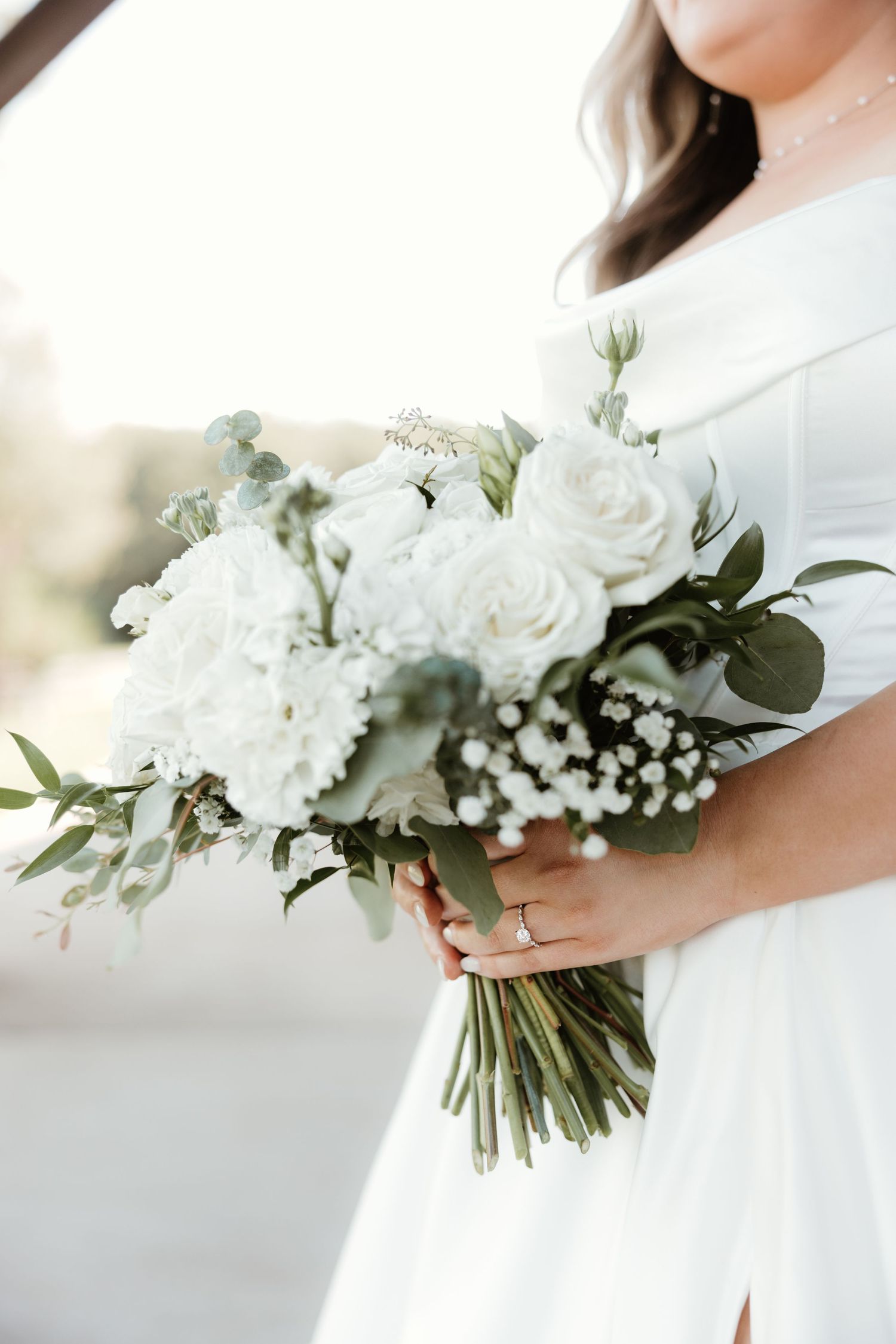A delicate white wedding bouquet featuring roses, baby's breath and eucalyptus held against a white dress.