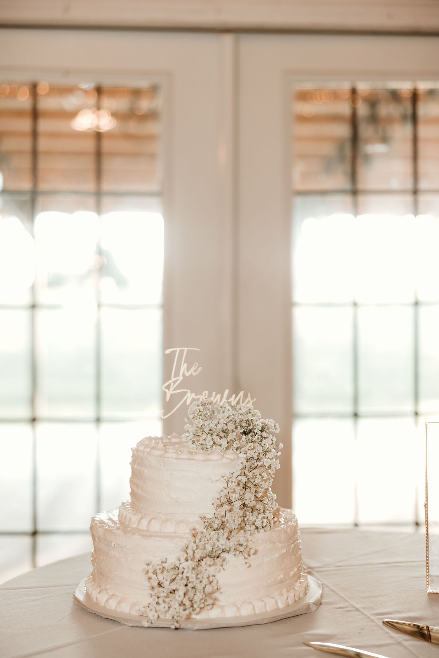 Elegant two-tier white wedding cake with cascading baby's breath flowers and 'The Browns' cake topper in an airy venue.