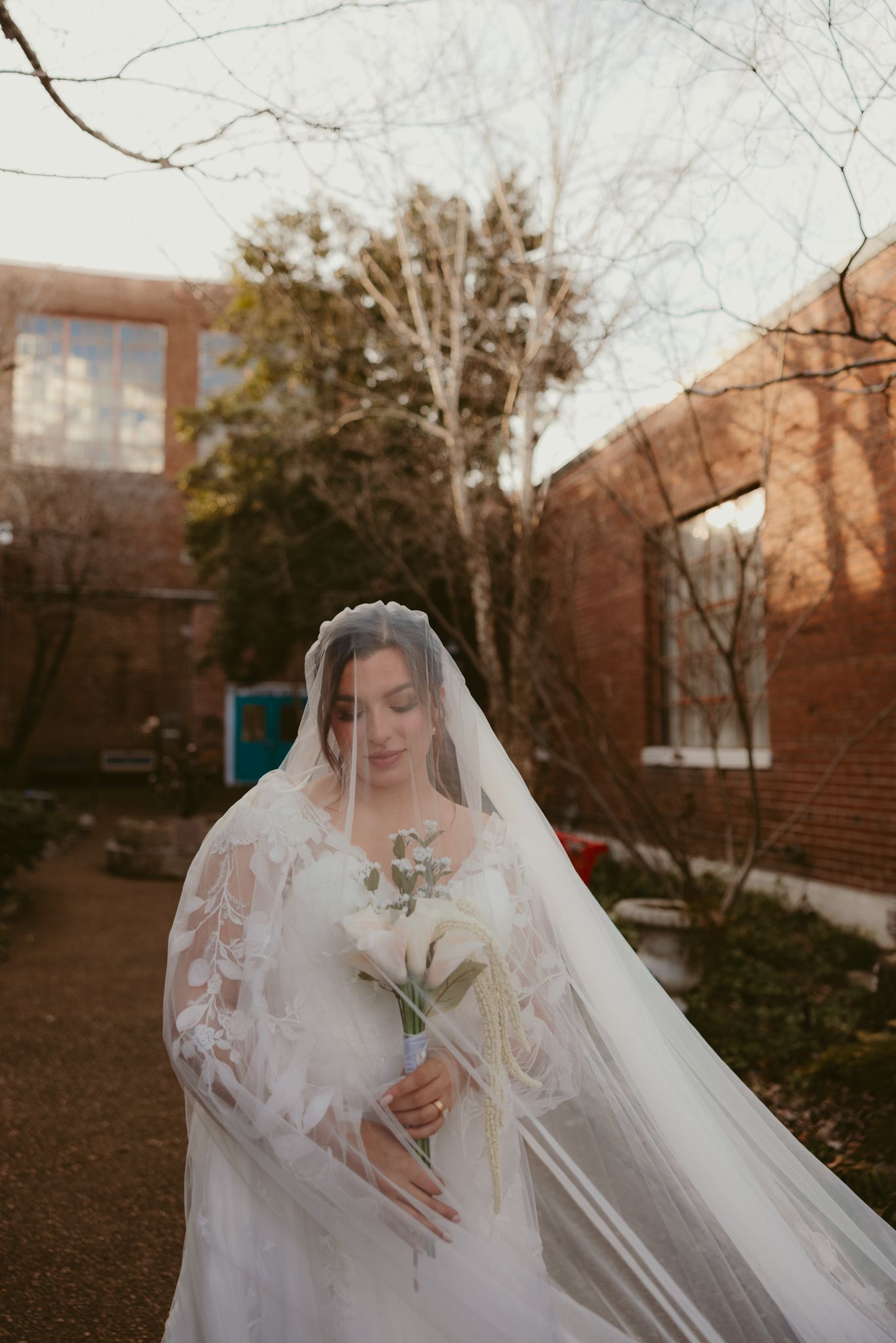 A bride in a lace gown holds a bouquet while her veil flows in the breeze near brick buildings.