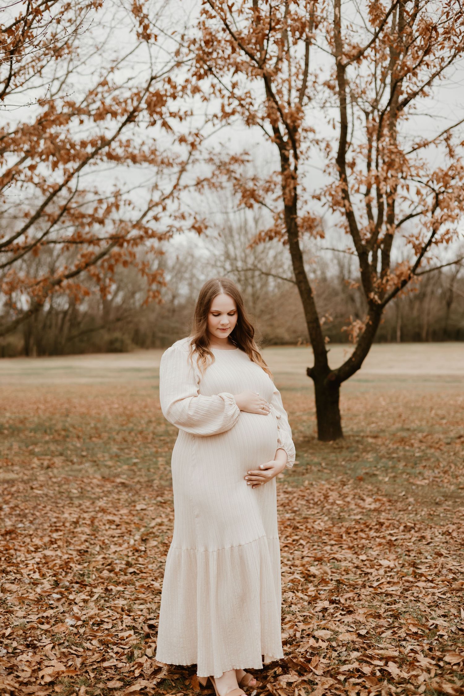 A pregnant woman in a white maxi dress poses in front of autumn trees.