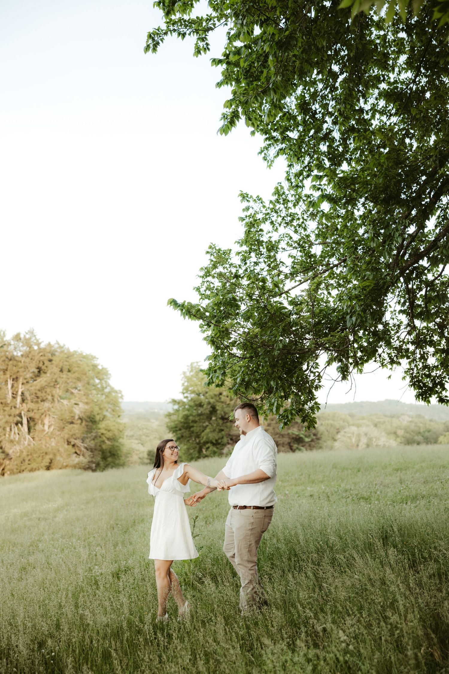 A couple stands together in a sunlit meadow surrounded by trees on a warm summer evening.