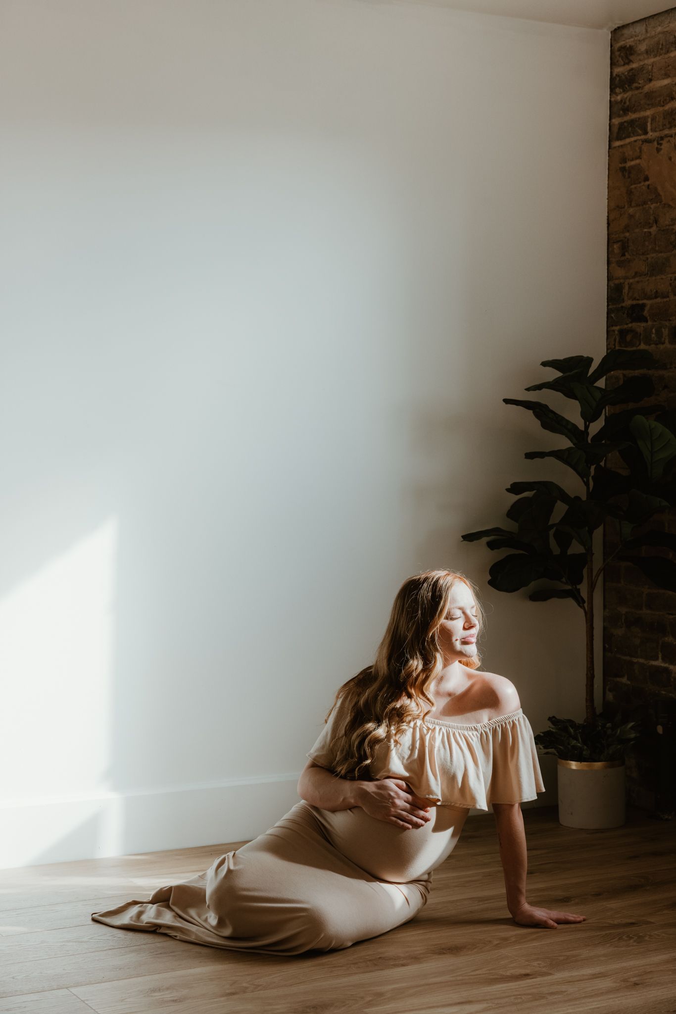 Person in off-shoulder white dress sits on wooden floor in sunlit room with potted plant and exposed brick wall.