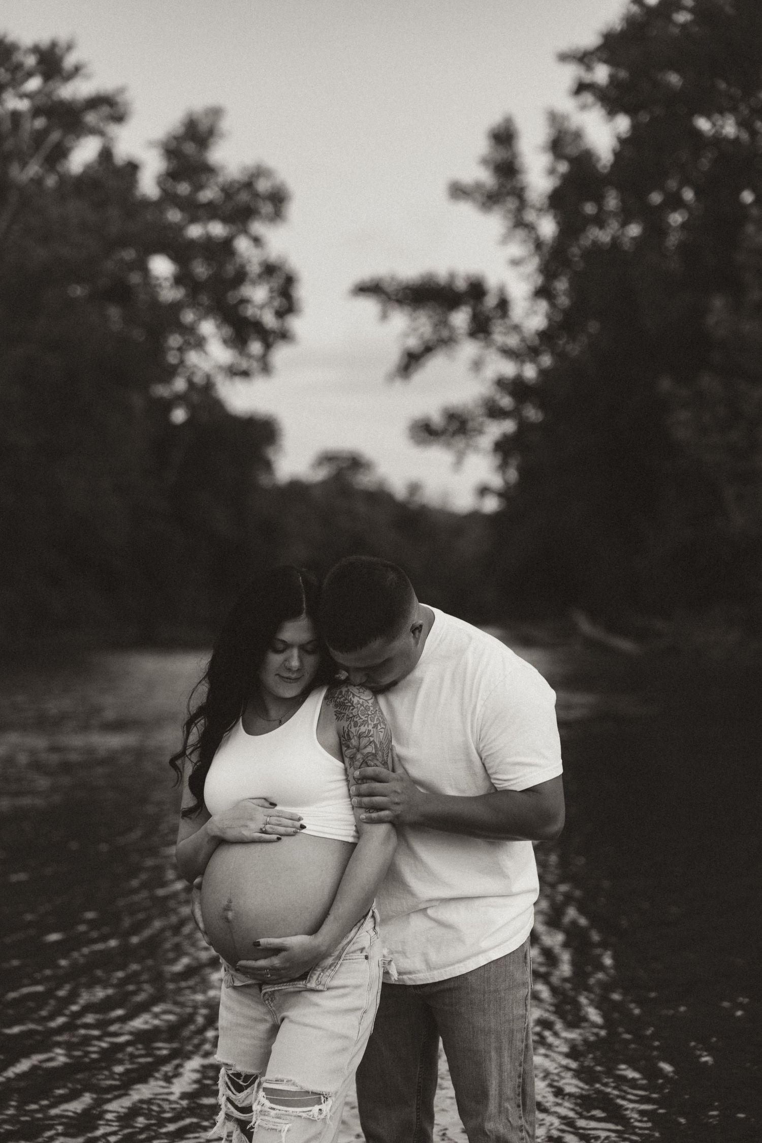 A couple in white shirts share an intimate moment outdoors by a lake at sunset in a black and white photograph.