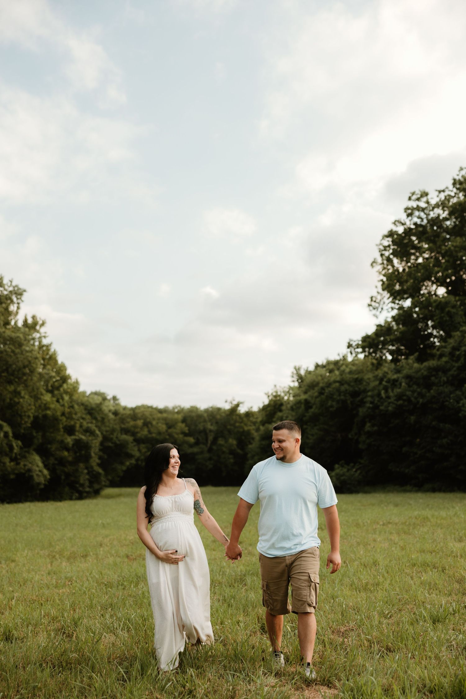 A couple walks hand in hand through a lush green meadow on a cloudy summer day.