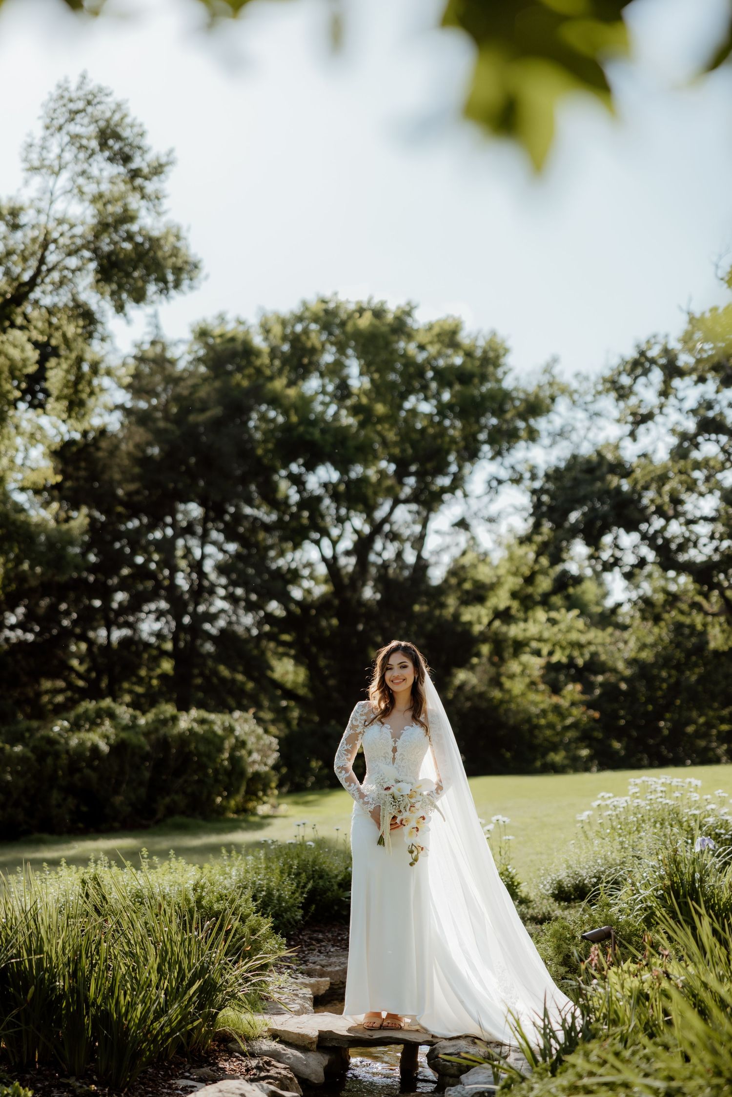 Bride in white lace long-sleeve gown with veil holds bouquet in lush green garden setting on sunny day.