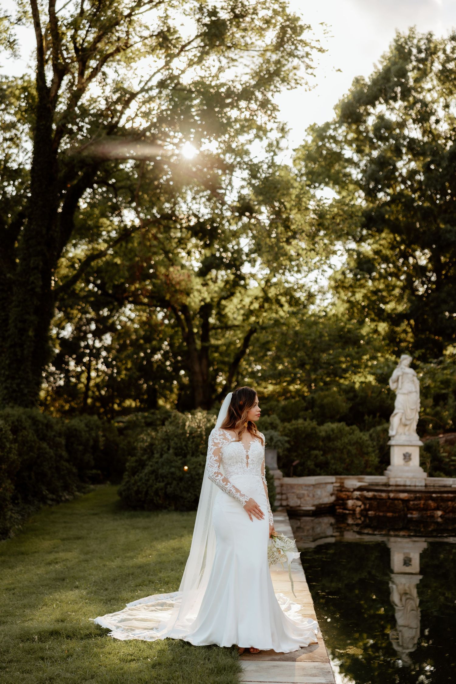 Bride in a long-sleeve lace wedding gown with veil stands in a sunlit garden near a stone statue.