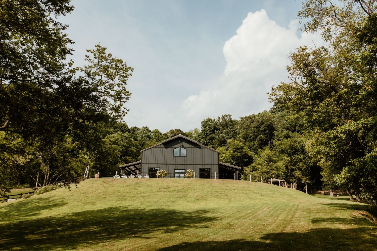 A gray barn-style modern home sits atop a manicured green hillside surrounded by tall trees on a sunny day.