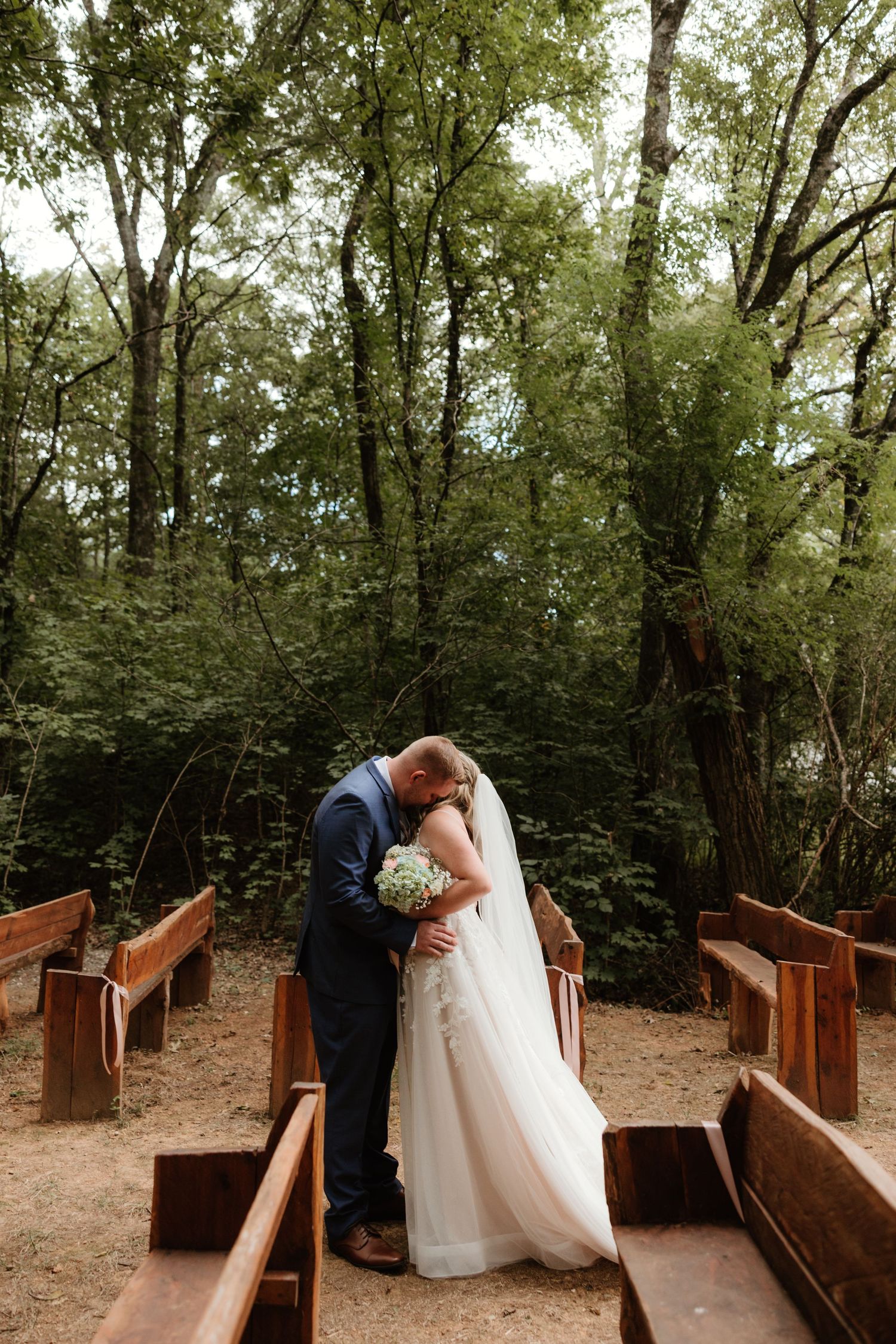 A tender moment shared between couples in a rustic wooden bridge walkway surrounded by lush green trees.
