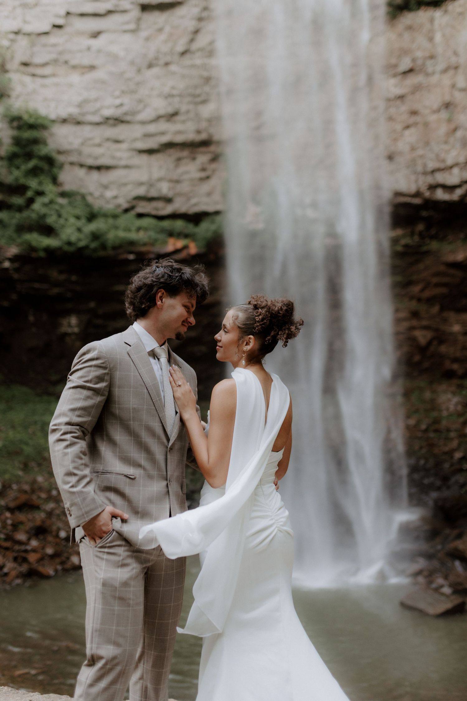 Bride and groom share an intimate moment in front of a cascading waterfall, bride wearing a flowing white cape gown.