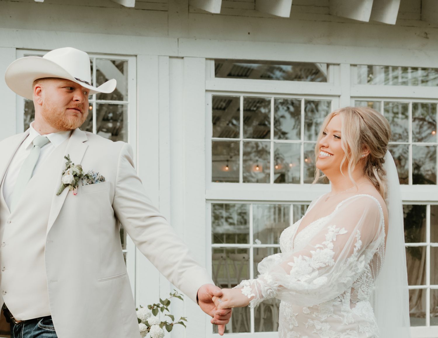 A couple shares a joyful moment in formal wedding attire outside a white building with windows.